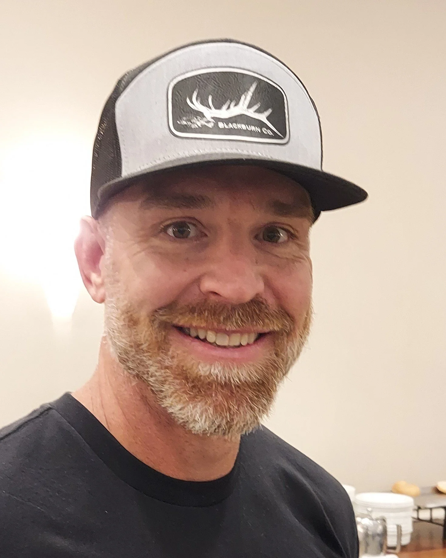 A smiling man with a beard wearing a gray and black trucker hat with a deer logo and a black t-shirt, standing indoors with a plain light-colored wall and some dishes in the background.