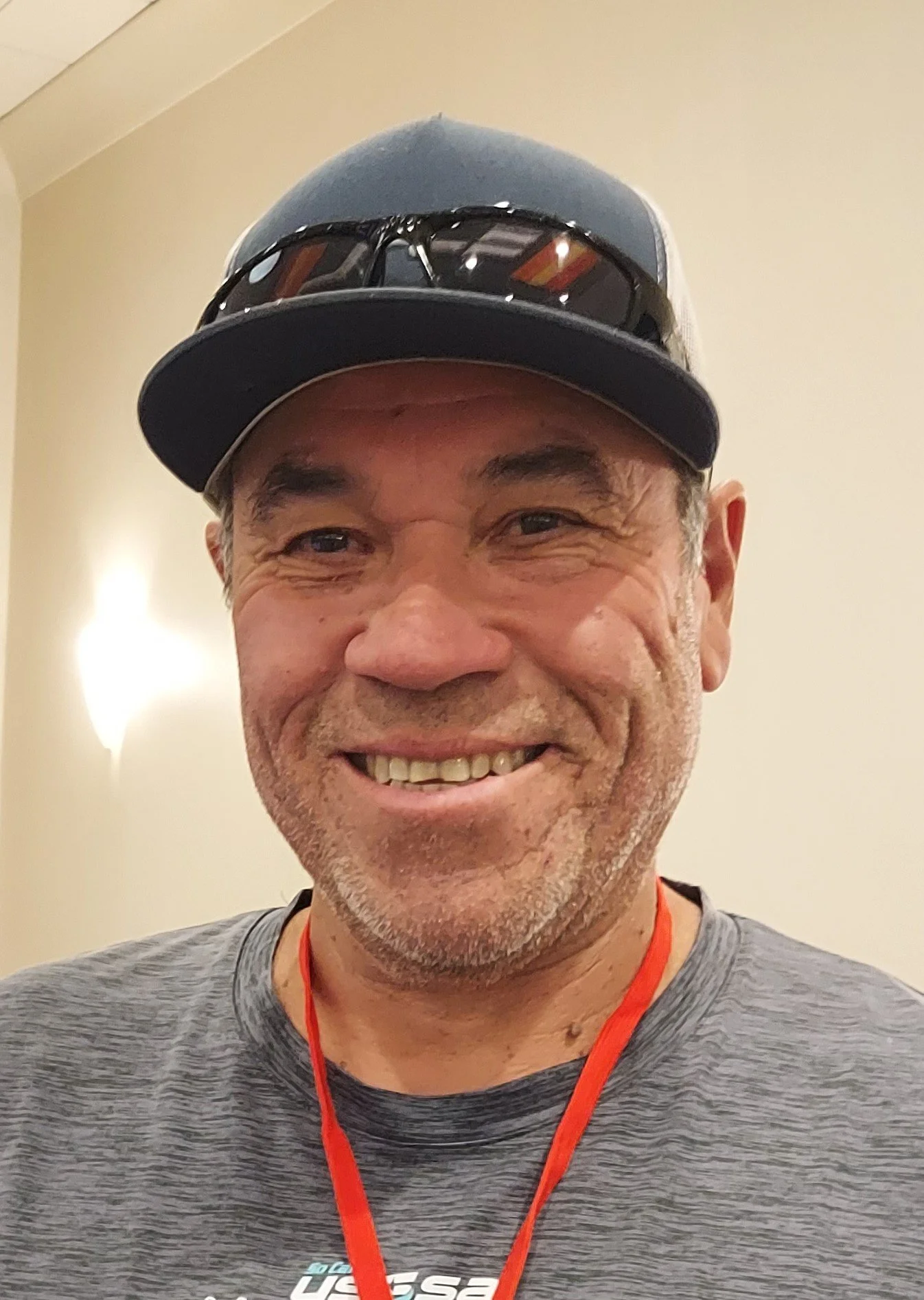 A smiling man wearing a baseball cap, sunglasses, a gray athletic shirt, and a red lanyard.