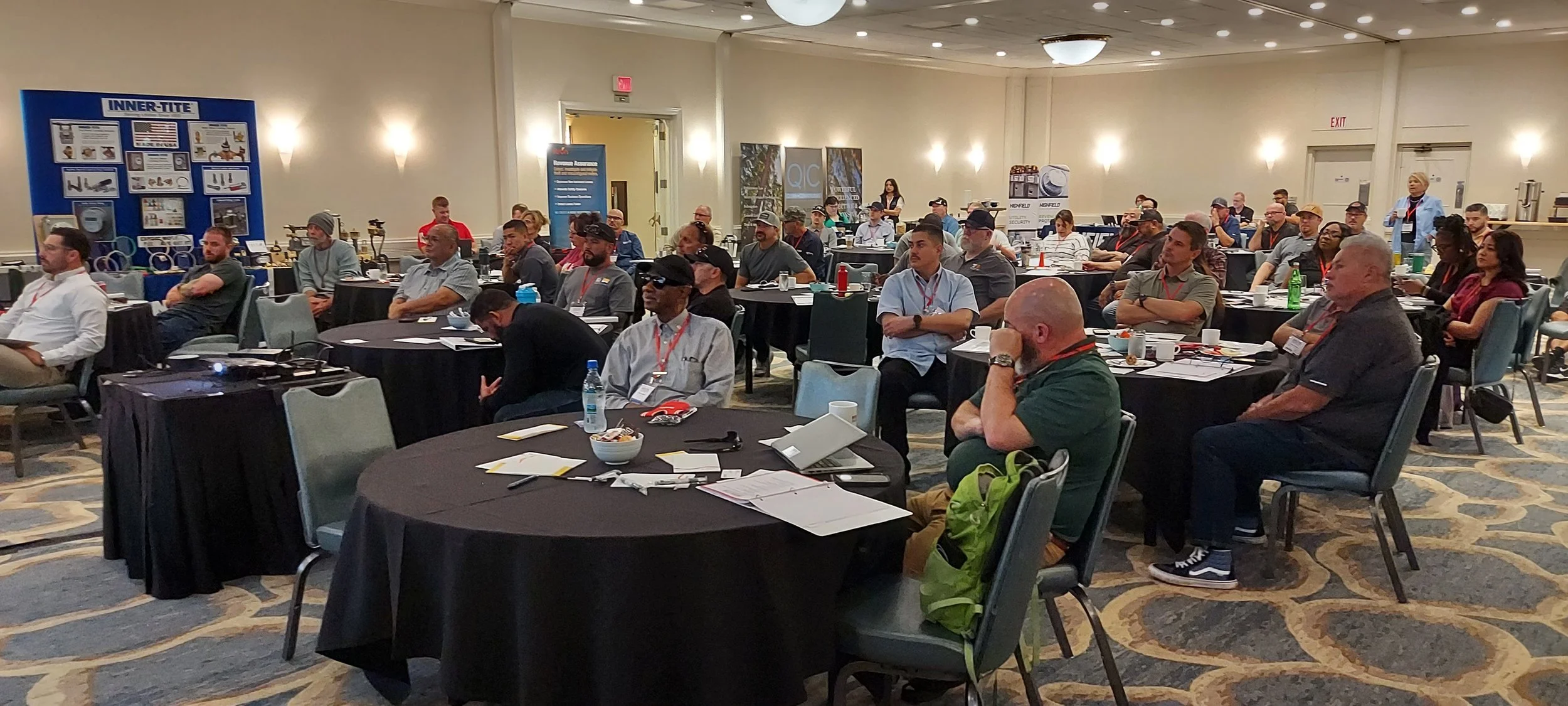 Conference room filled with attendees sitting at round tables, listening to a presentation or speaker, with some taking notes. The room has beige walls, carpeted floor, and booths with informational displays in the background.