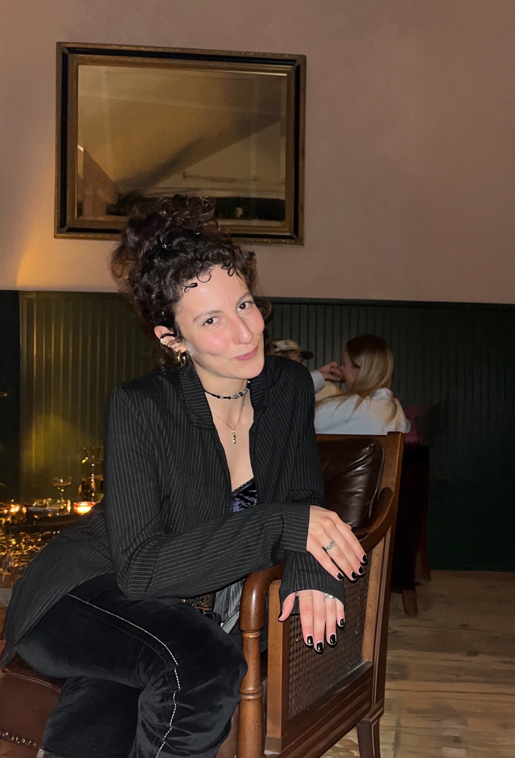 A Los Angeles based astrology and tarot readr with dark, curly hair styled in a messy bun, wearing a black pinstripe blazer and black pants, sitting on a wooden chair in a dimly lit event space with other patrons and a mirror in the background.