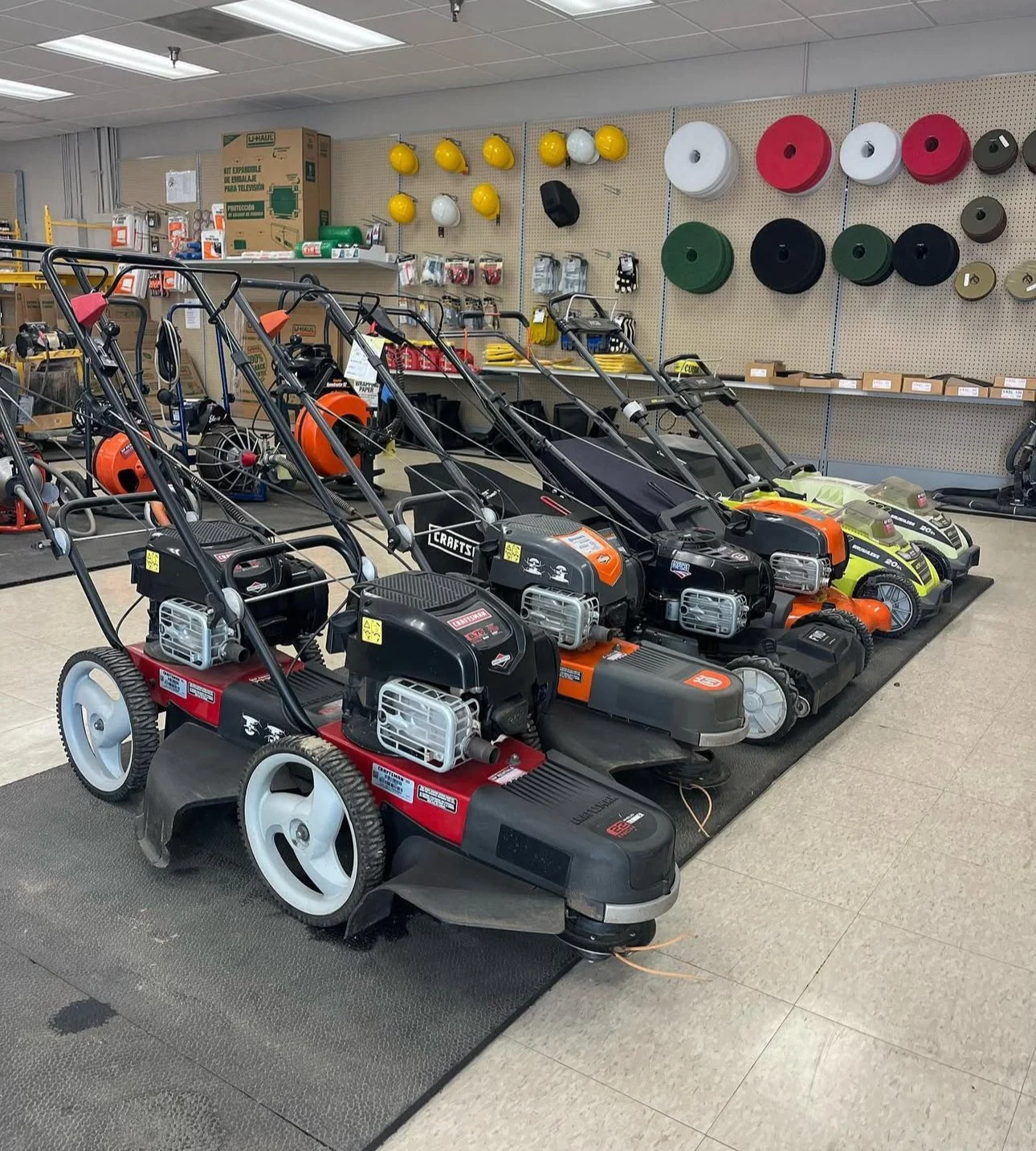 Several lawn mowers and outdoor power equipment displayed in a store aisle with various safety helmets and hardware on the wall behind them.