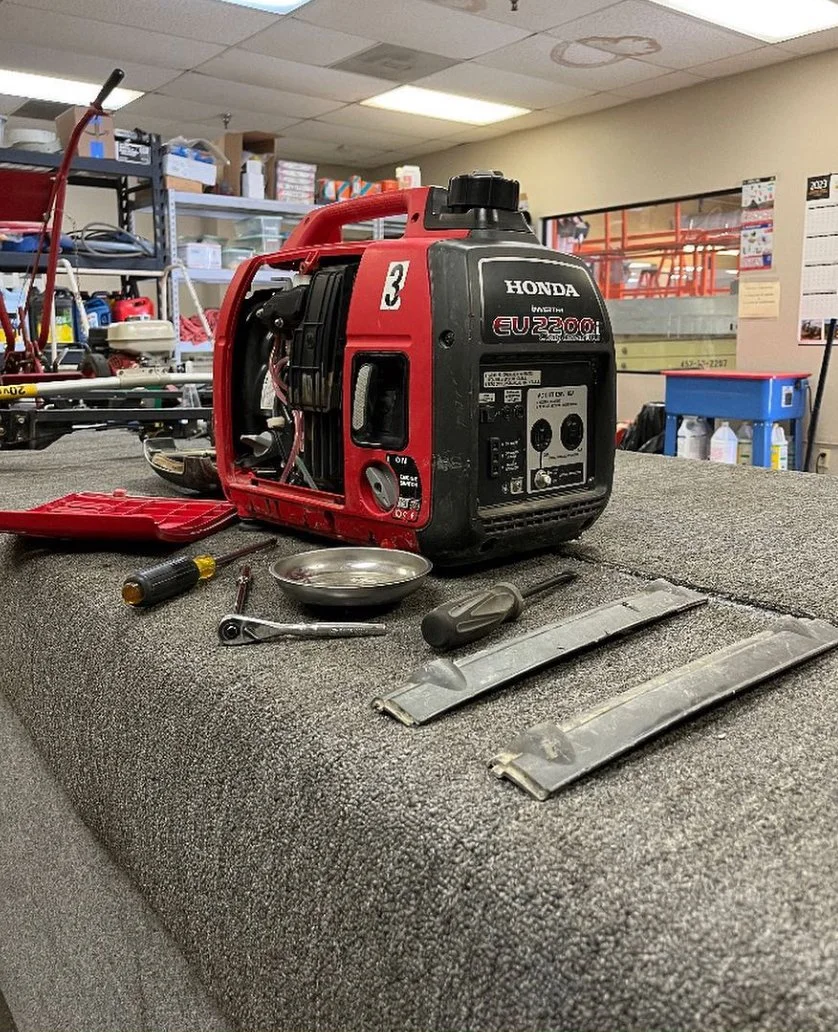 A disassembled portable Honda EU2200i generator on a workbench, surrounded by tools including screwdrivers, a ratchet, and metal strips, in a workshop or repair room with shelves and equipment in the background.