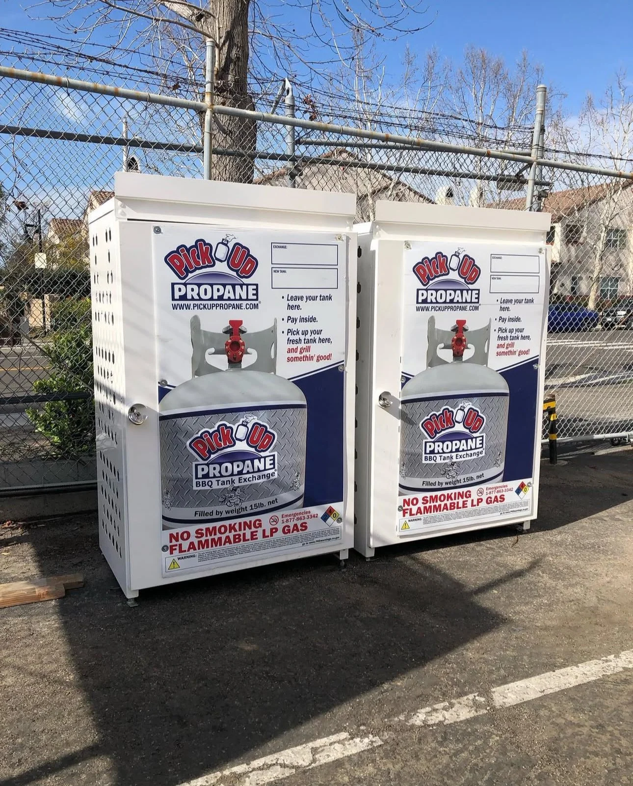 Two propane tank exchange lockers with instructions, warning labels, and branding for Pick Up Propane, located outdoors on a paved lot with a chain-link fence and leafless trees in the background.