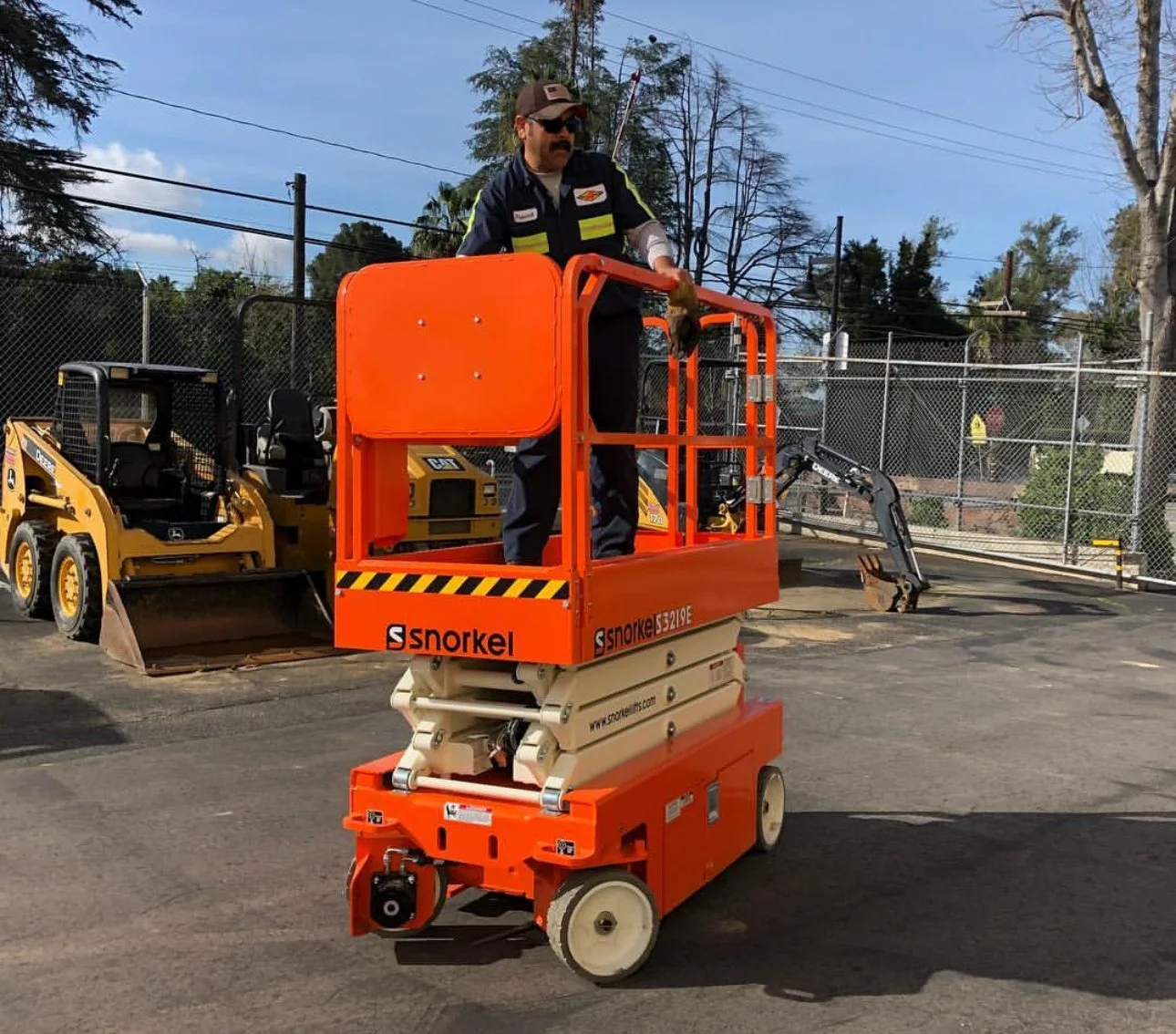 Person operating a bright orange Snorkel lift in a parking lot with construction equipment and fenced area in the background.
