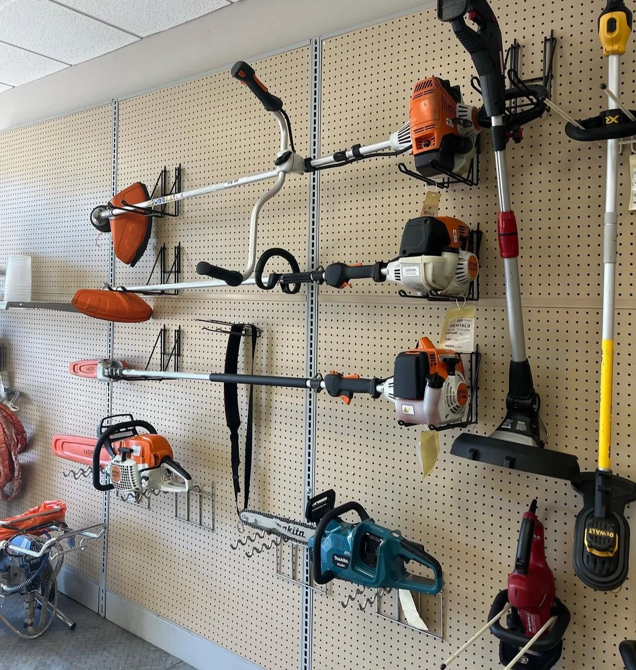 Wall-mounted display of various outdoor power tools including string trimmers, chainsaw, leaf blower, and pole saw in a hardware store.