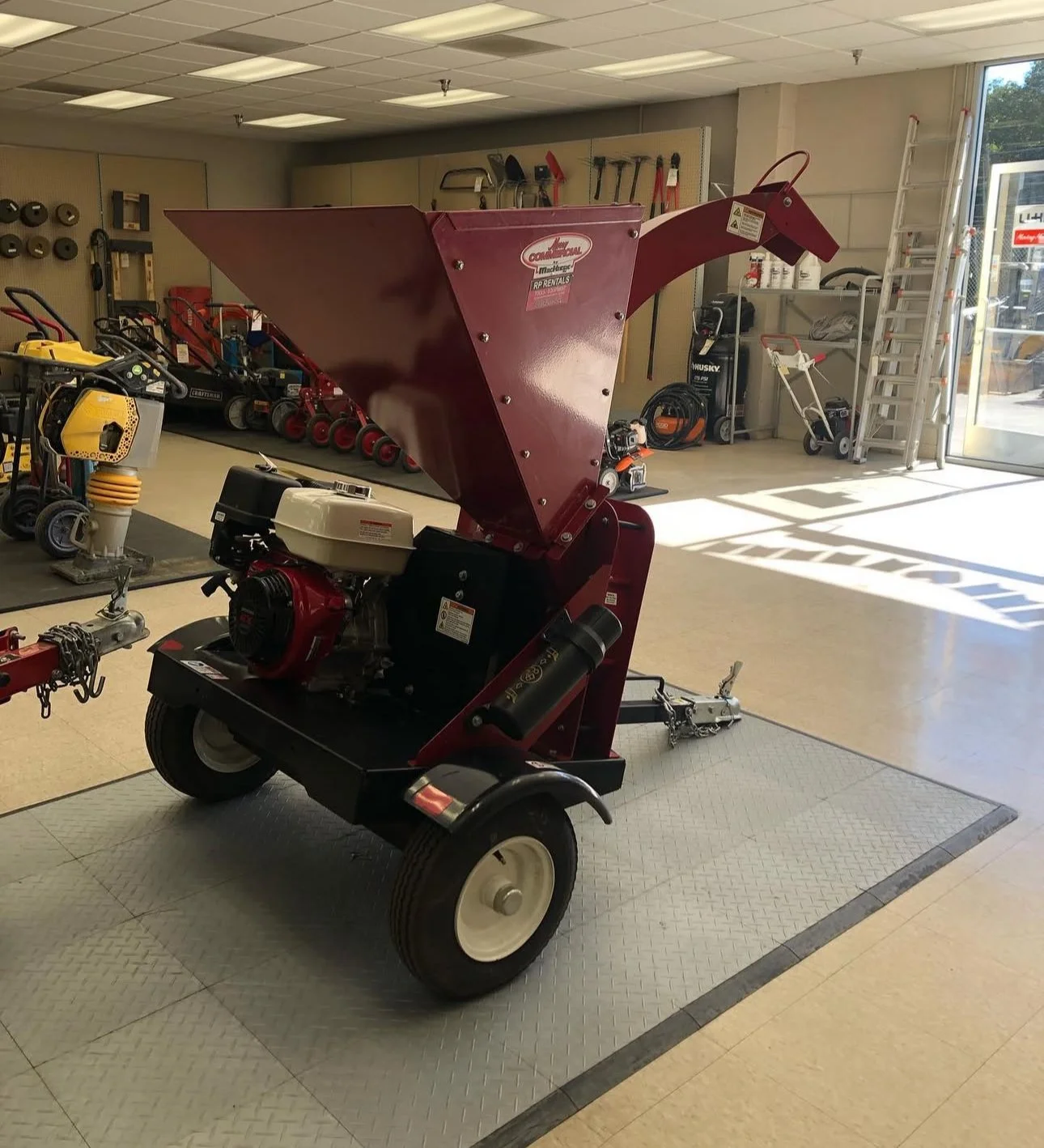 Red wood chipper with a large hopper, situated on an indoor showroom floor with various tools and equipment in the background.