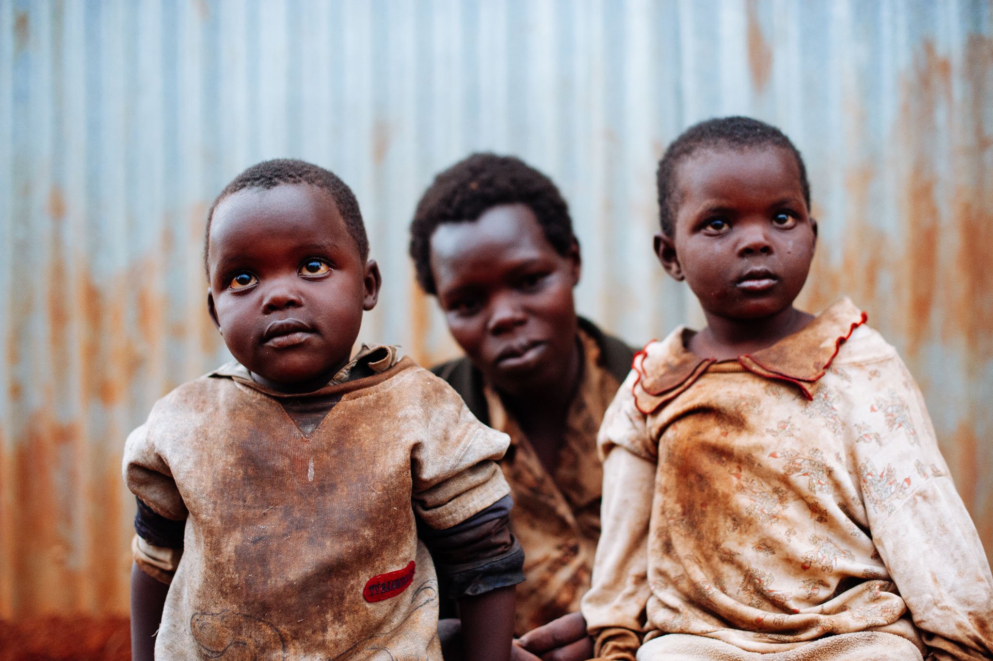 Two young children and a woman sitting outside in front of a corrugated metal wall, with a somber expression.