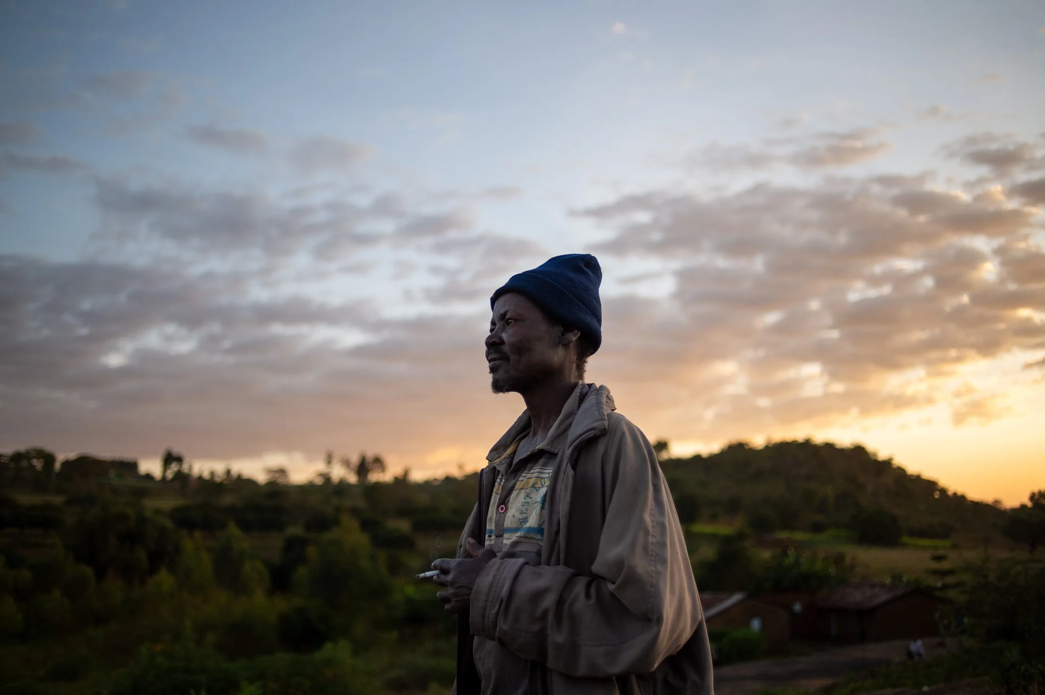 A man standing outdoors during sunset, wearing a blue beanie and a gray jacket, looking to the side with a scenic landscape and partly cloudy sky in the background. Turbo, Kenya