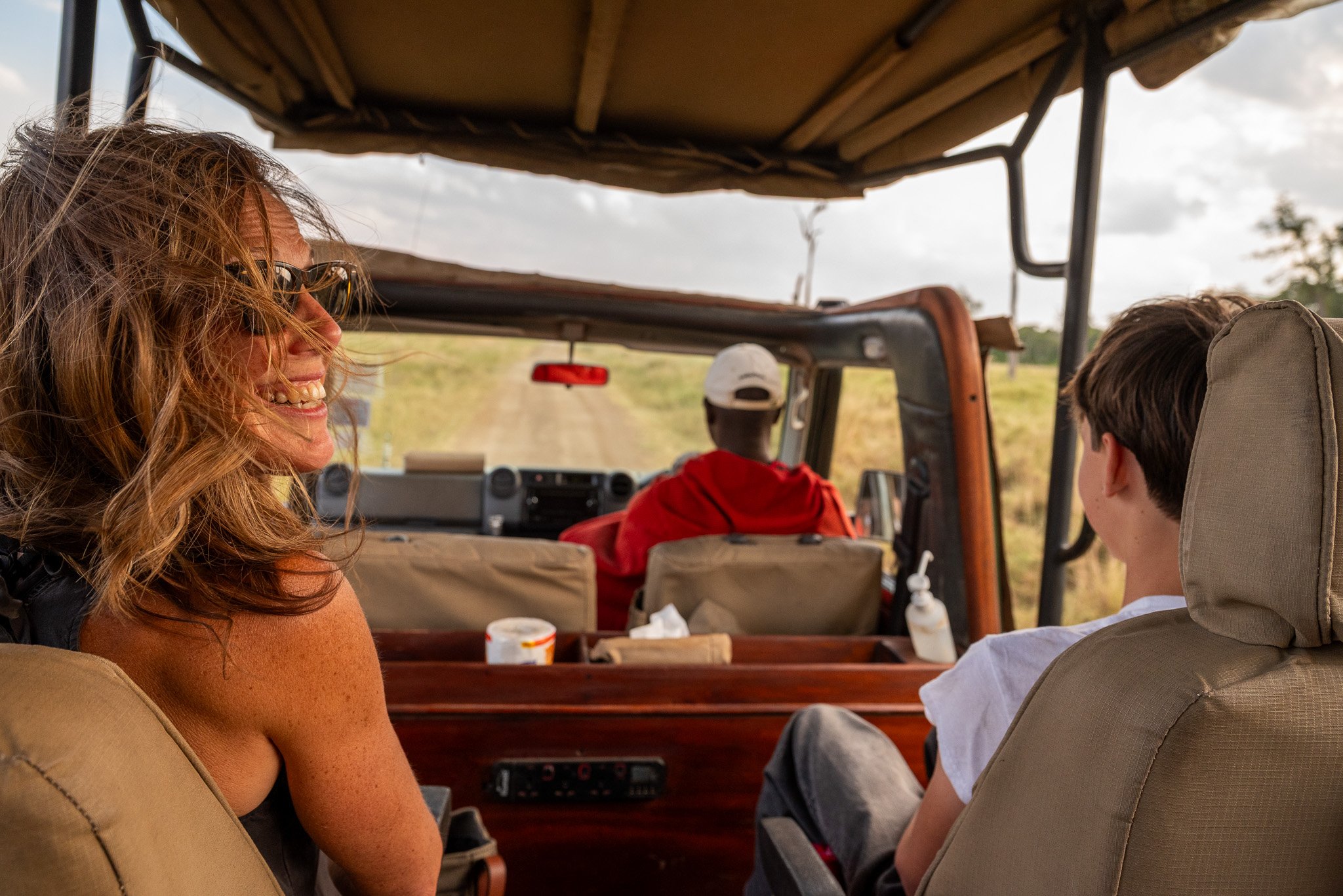People riding in an open-air safari vehicle through a rural area, with a woman smiling and looking to the side, and three other travelers, including a young boy, all seated inside the vehicle.