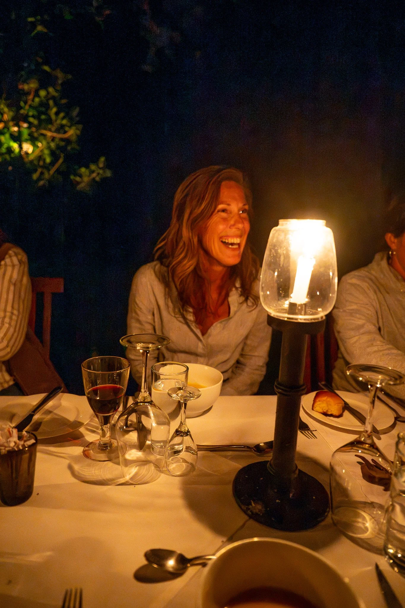A woman with long, wavy hair laughing during a dinner at night. She is sitting at a table with various glassware, a bowl, and a plate with bread. There is a tall lamp providing warm light on the table, and dark foliage or trees in the background.