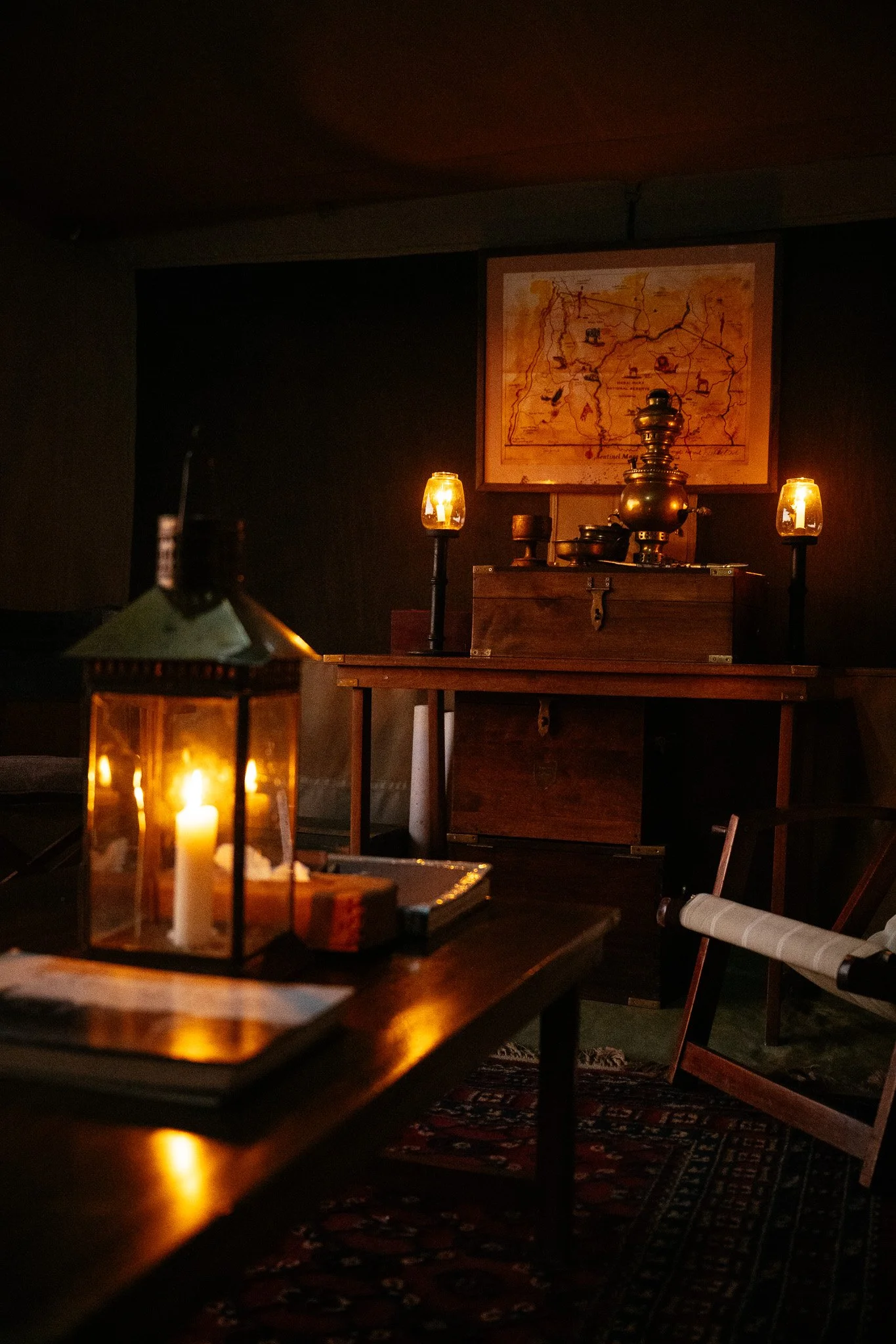 Dimly lit room with candles on tables, a framed map on the wall, and vintage lanterns, creating a cozy, rustic atmosphere.
