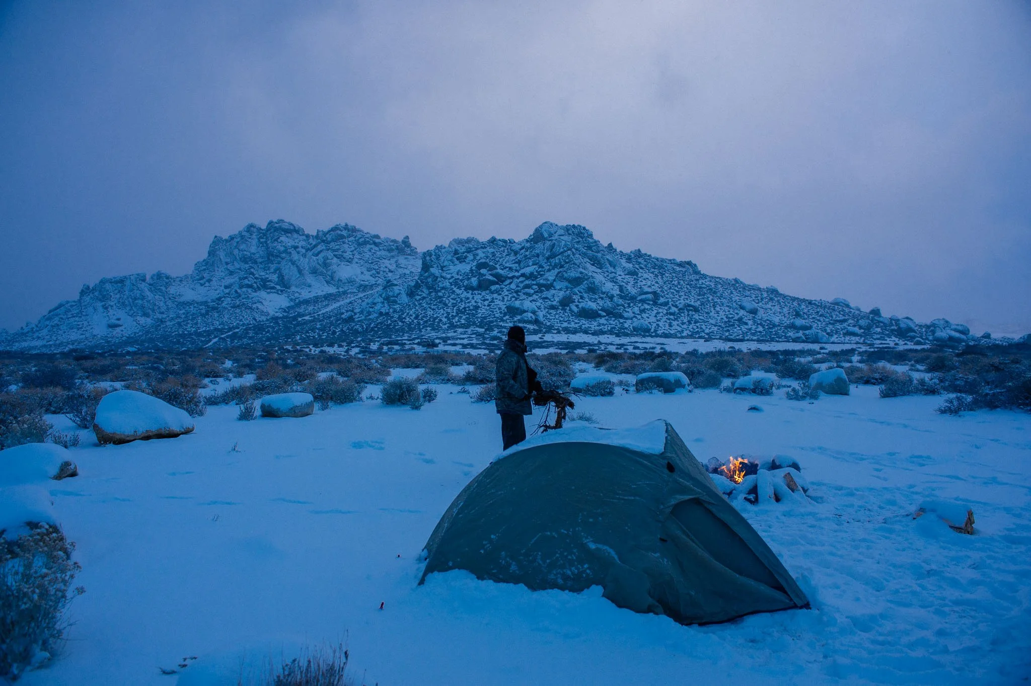 A person in outdoor winter gear standing near a tent in a snowy landscape with large boulders and a mountain in the background at dusk or dawn.