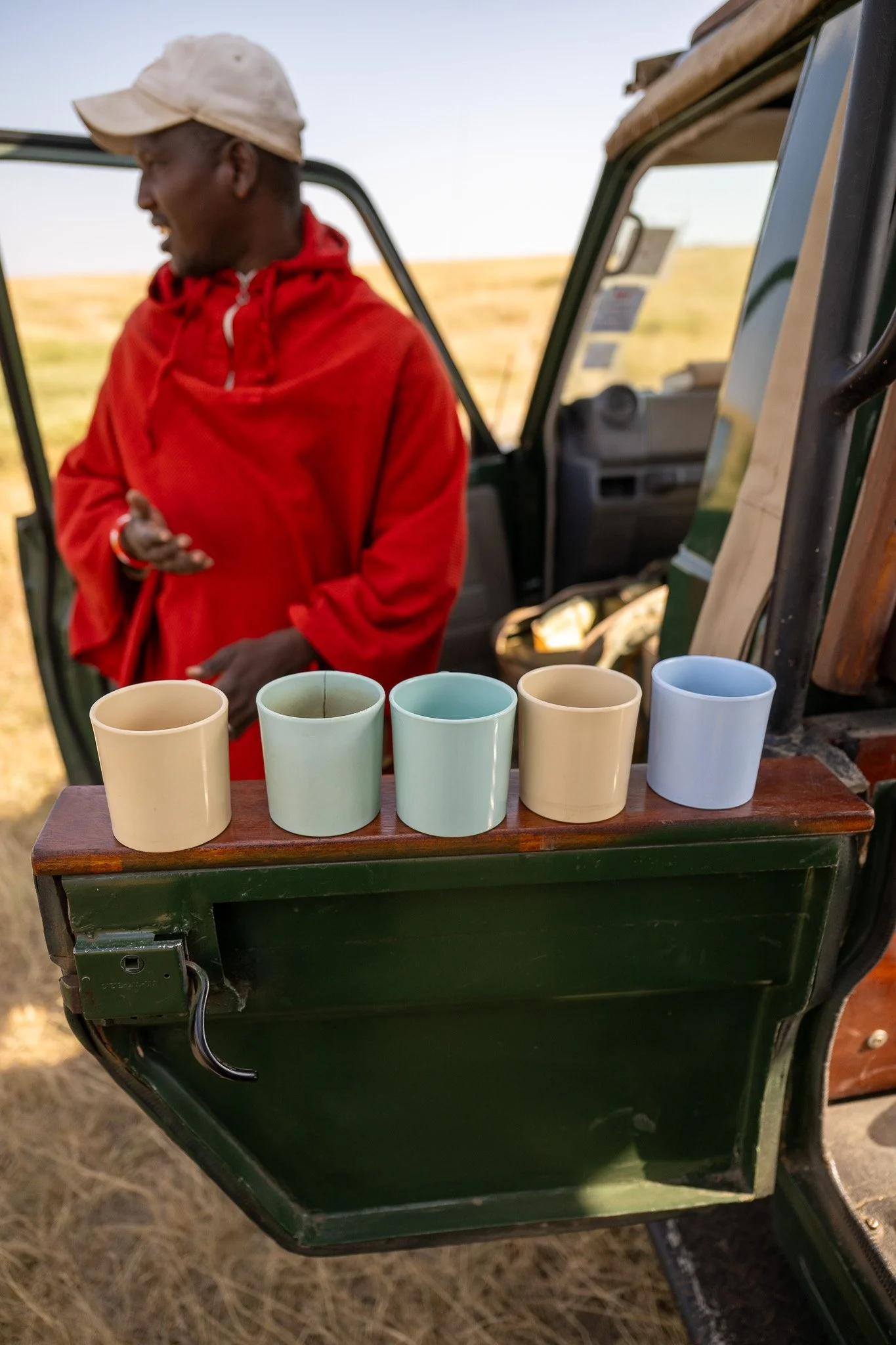 A man in a red hoodie and beige cap standing beside a green utility vehicle with white and blue cups arranged on a wooden surface, in an outdoor setting with dry grass and a clear sky.