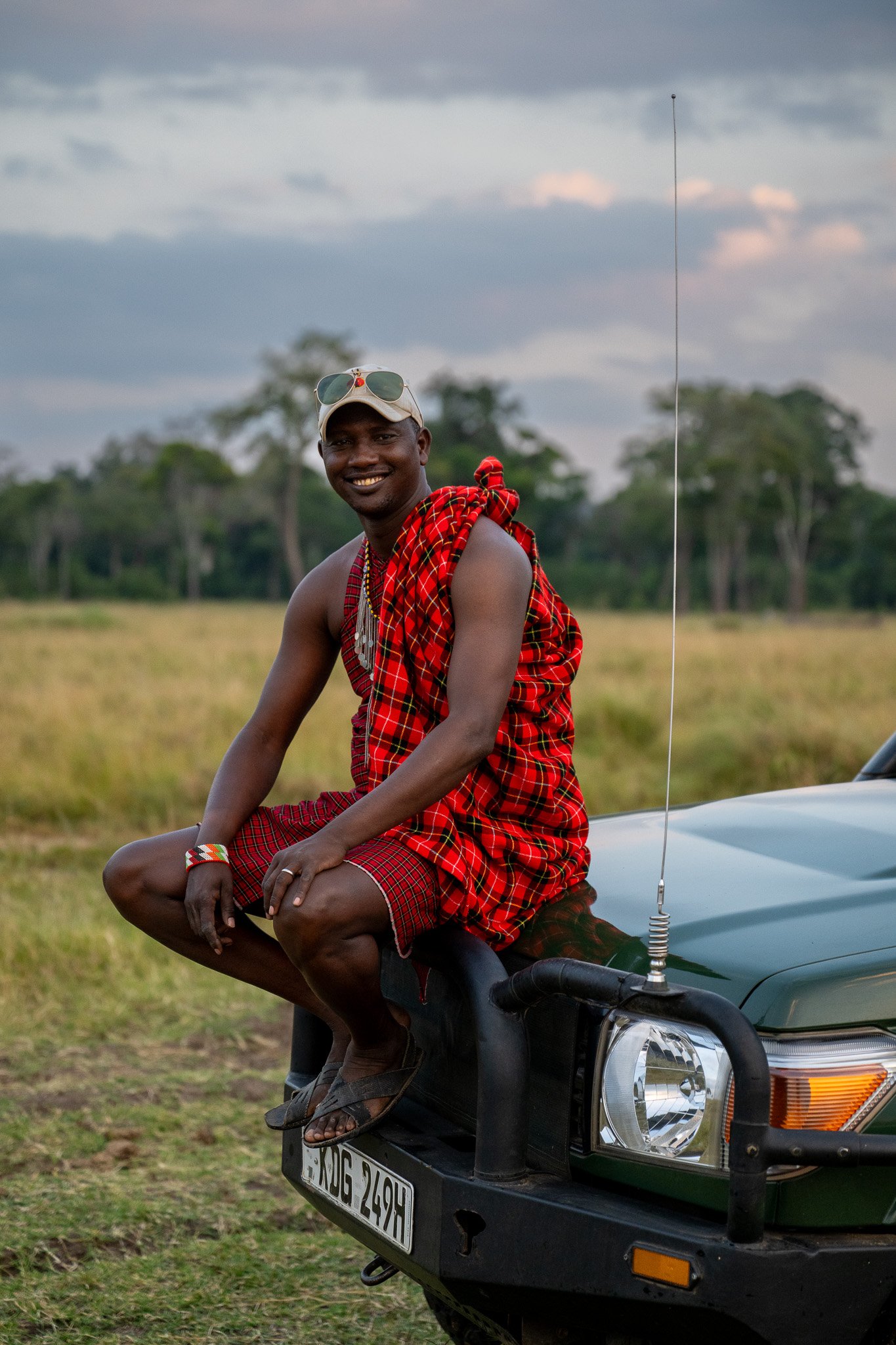 A man with dark skin, smiling, dressed in traditional Maasai clothing, and wearing sunglasses on his cap, sitting on the hood of a black vehicle with a rural landscape and trees in the background.