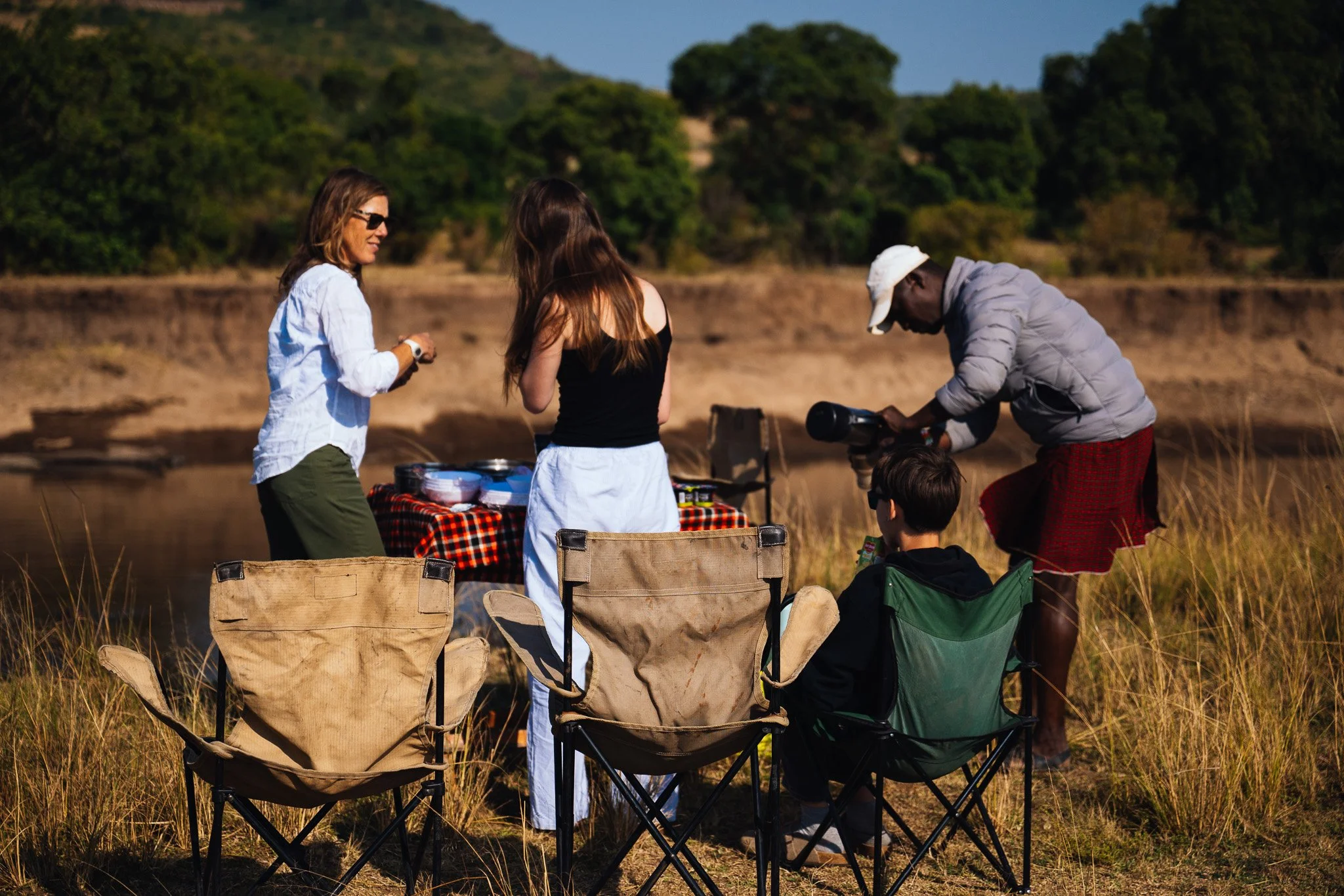 Group of people outdoor near a lake with folding chairs and various items on a checkered tablecloth, surrounded by trees and hills.