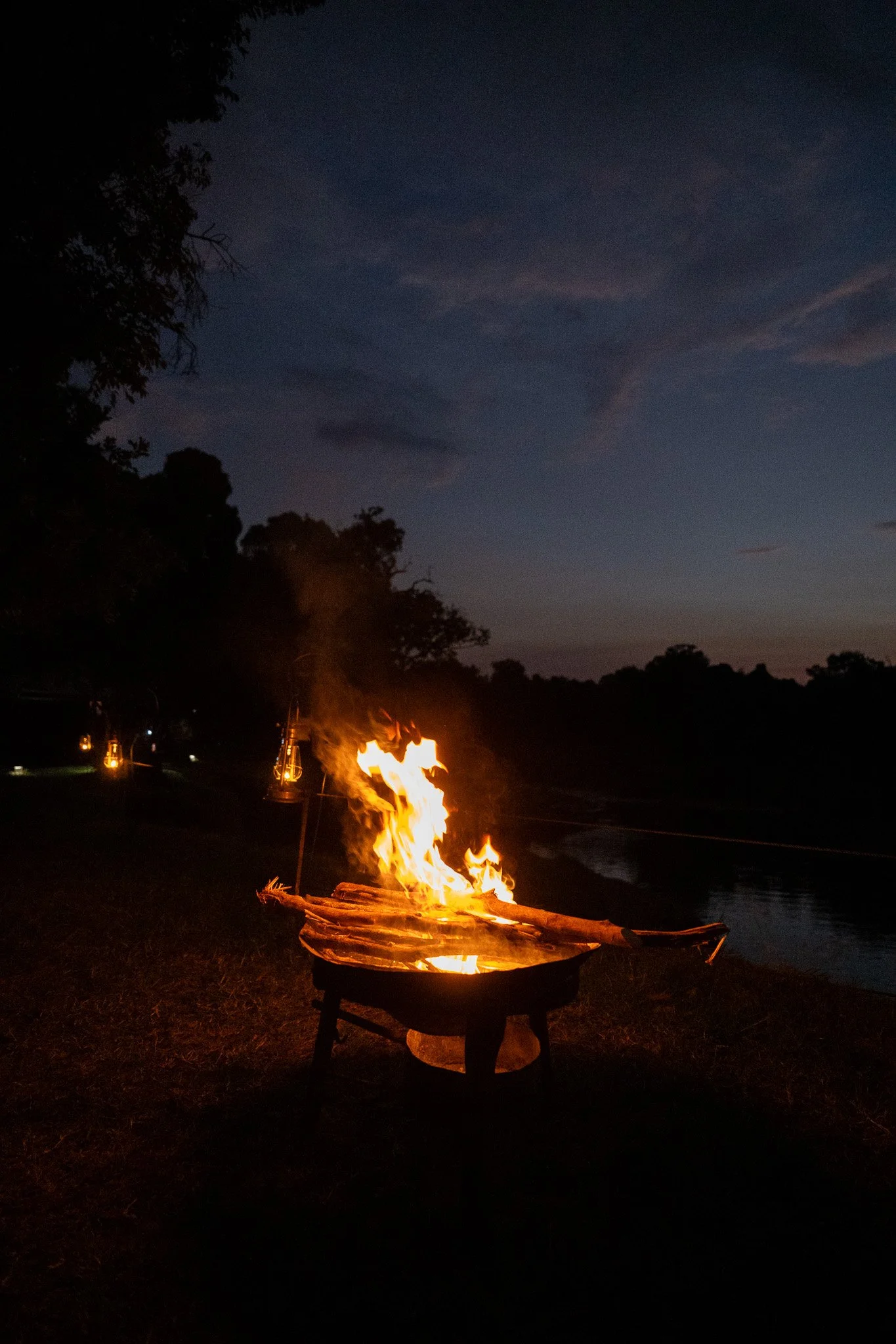 Fire burning in a metal fire pit at night near a body of water, with trees and lanterns in the background and a dark sky overhead.