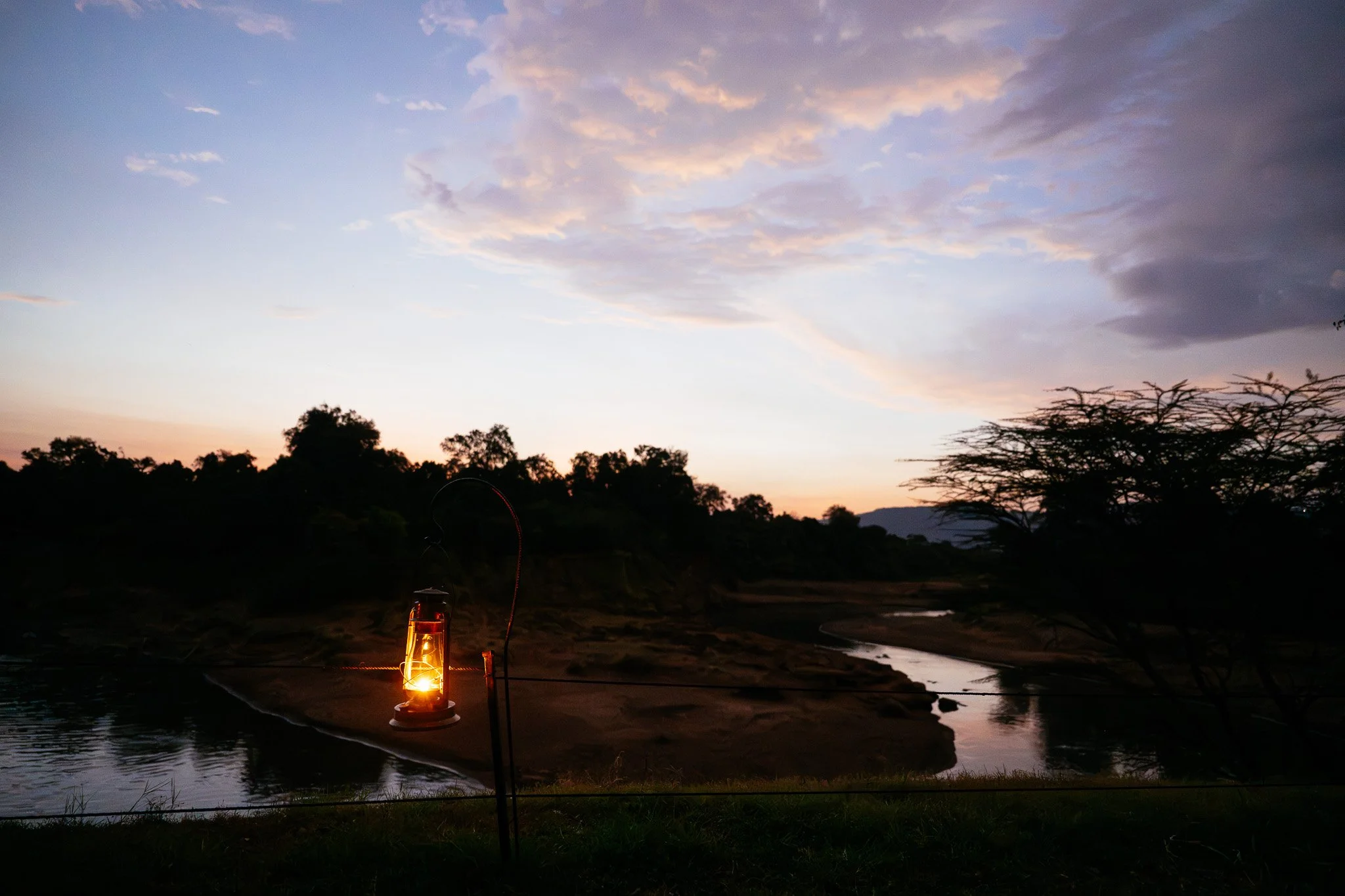 Sunset over a river with trees in the background, a lantern with a candle inside placed on the ground near the riverbank, and a fishing rod leaning against it.