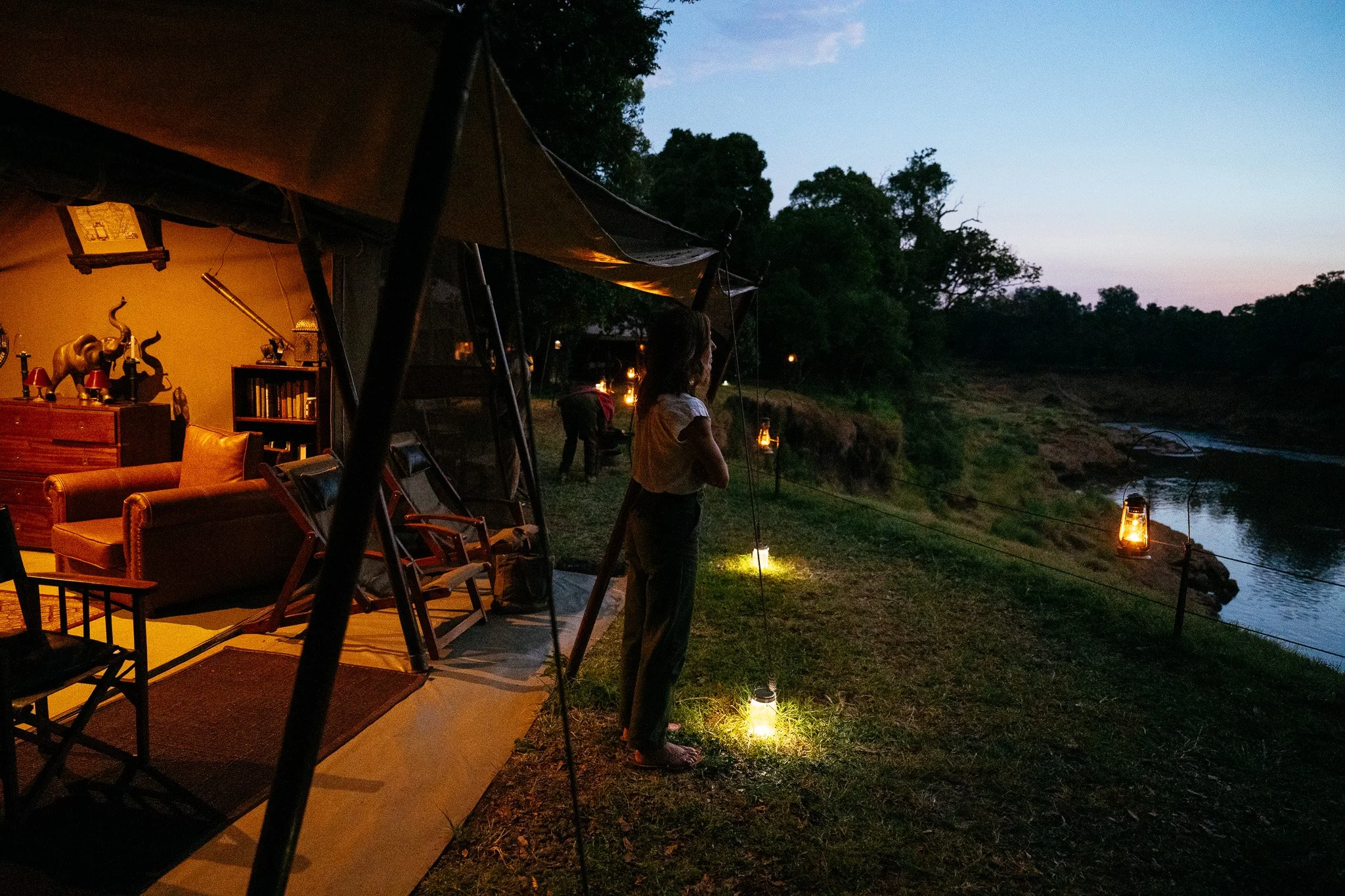 Outdoor evening scene with a woman by a river, lanterns illuminating the area, and a tent with the interior showing furniture and decorations, surrounded by trees.