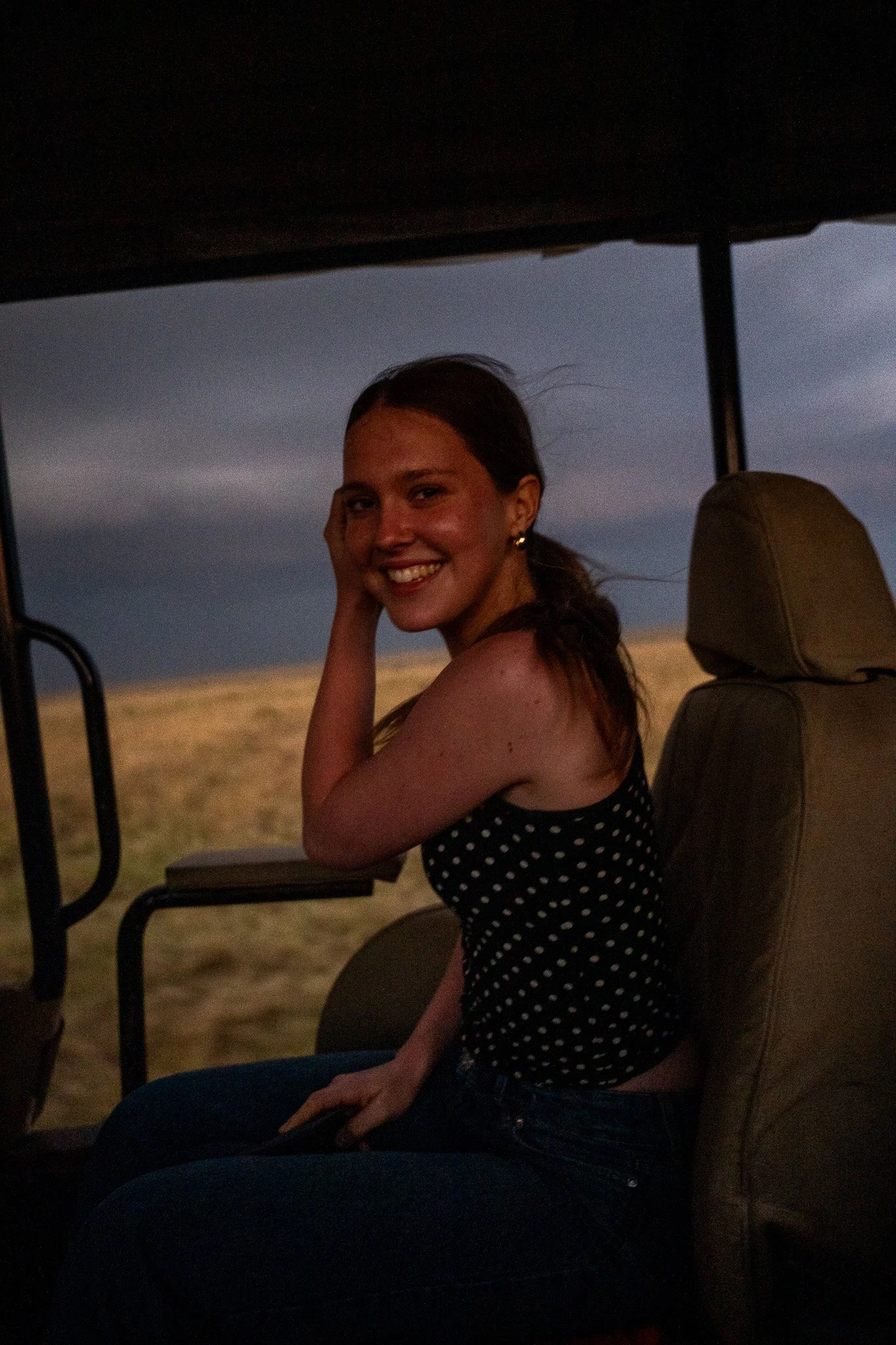 A woman with dark hair smiling while sitting in a safari vehicle at sunset, wearing a polka dot top and earrings.
