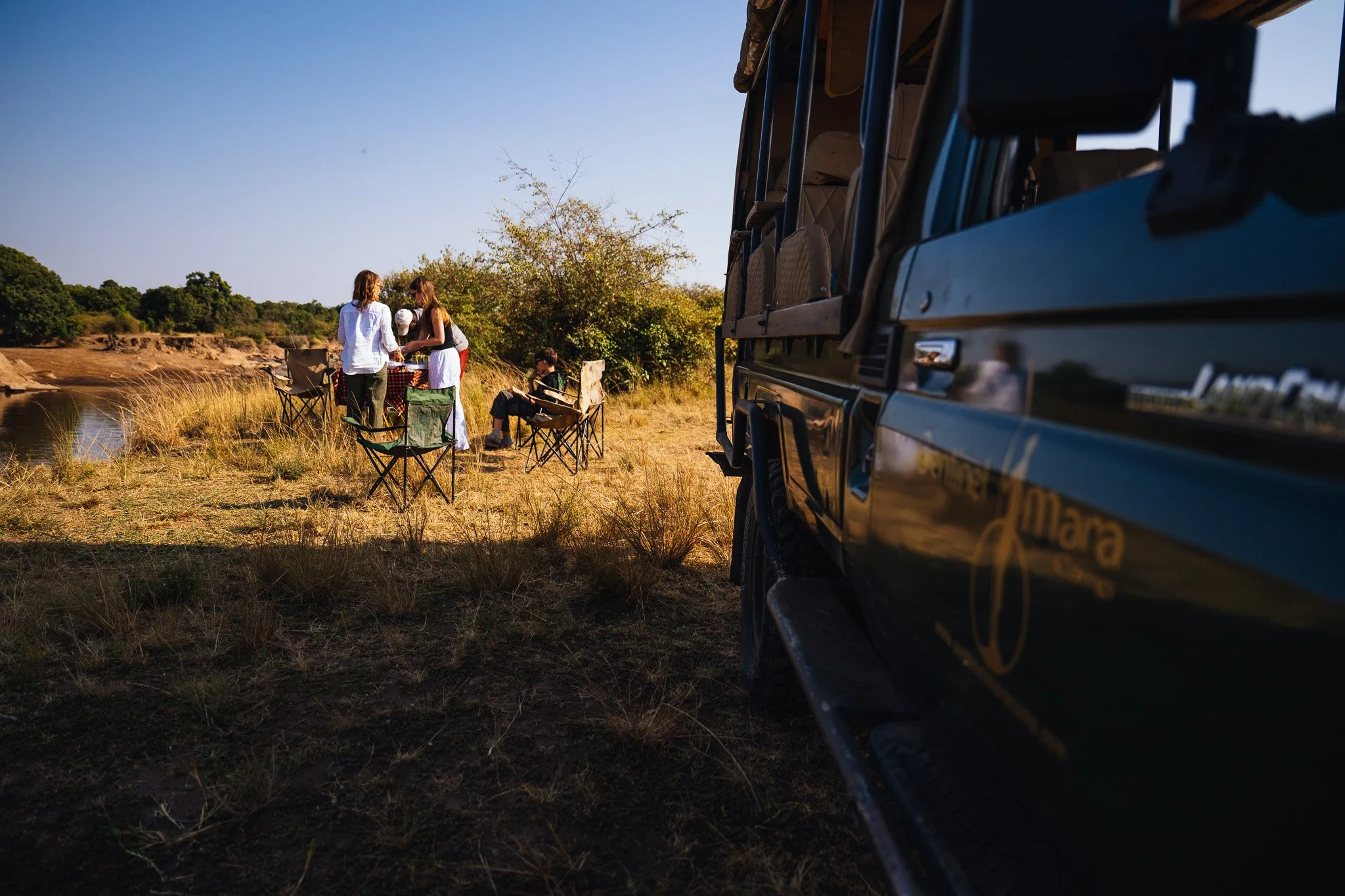 People gathered around a table outdoors by a river, with a vehicle parked nearby, in a rural natural setting with trees and grass during sunset.