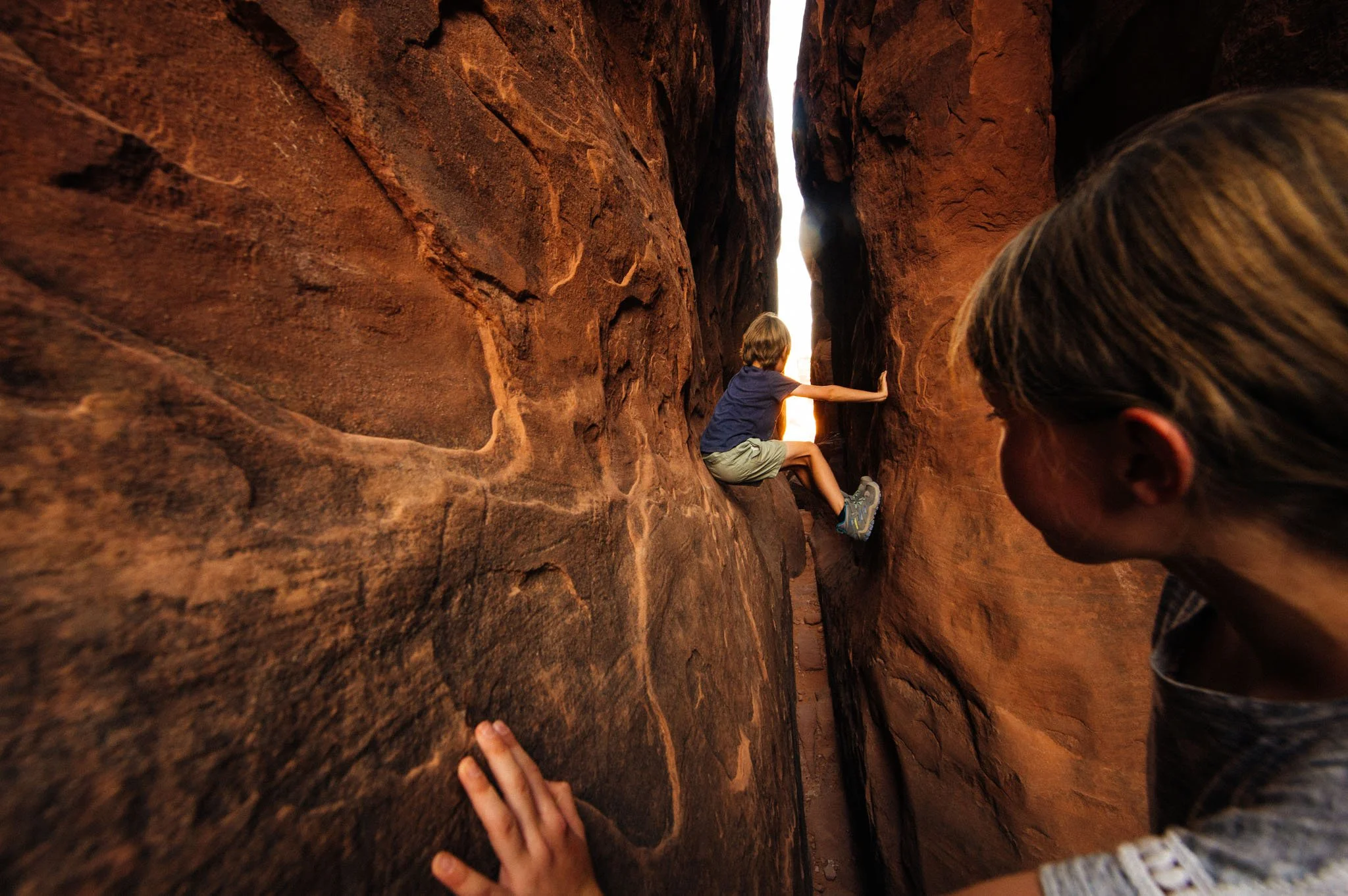 Two children are exploring a narrow red rock canyon, with one climbing and another observing from outside.
