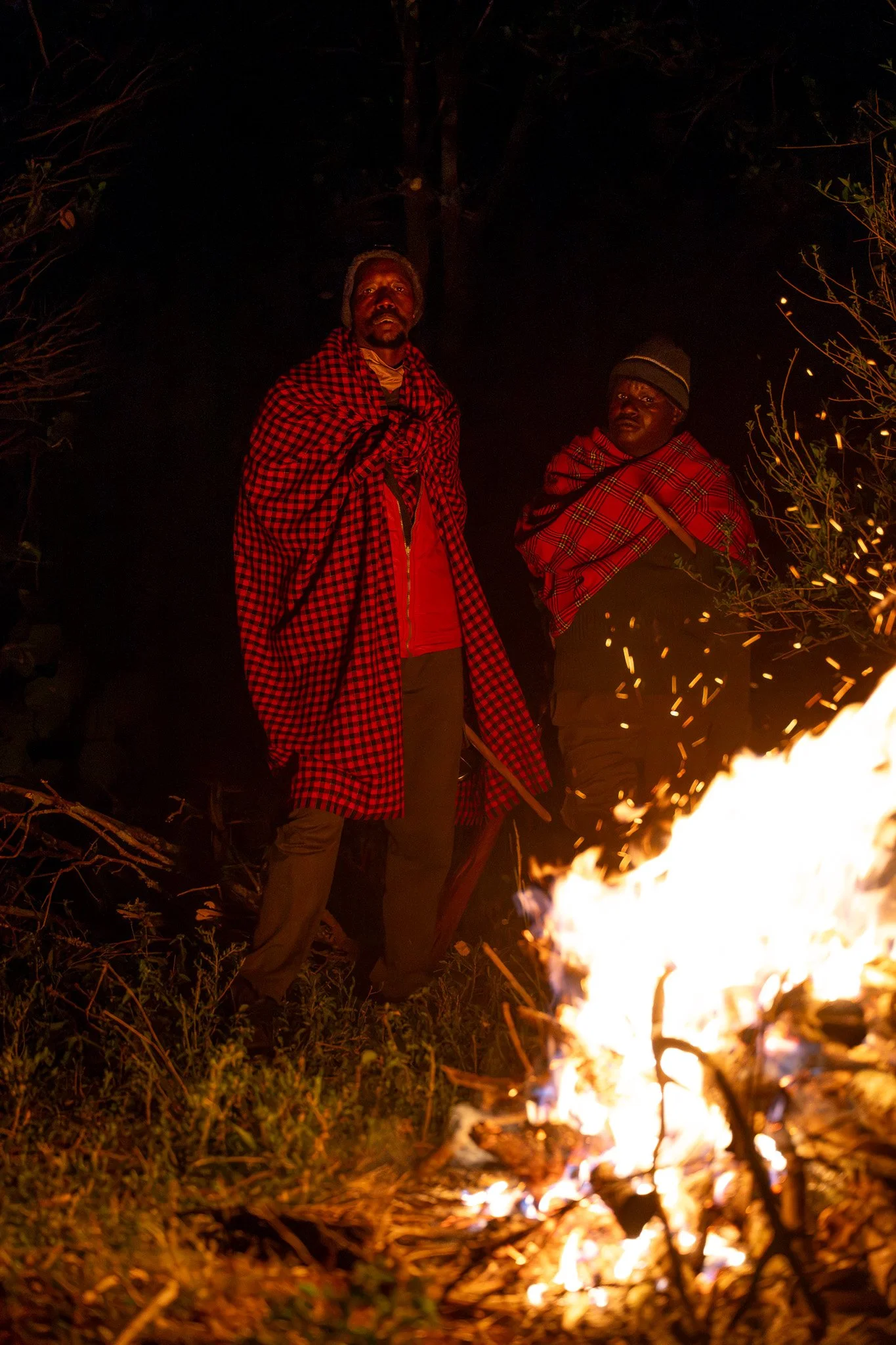 Two men standing near a fire at night, wearing traditional Maasai clothing and headgear.