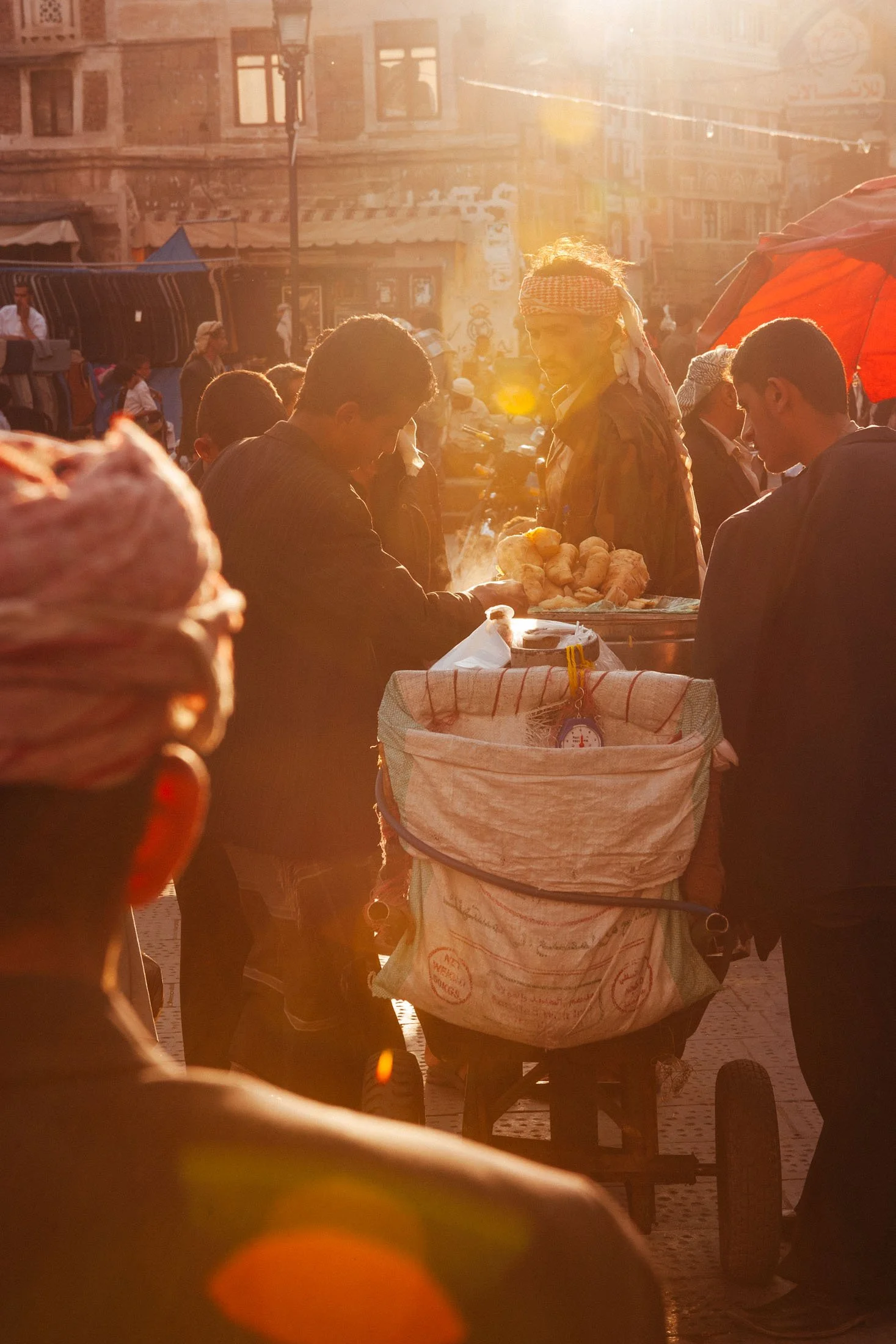 Street scene at sunset with a man buying bread from a vendor at a busy market, with people, buildings, and a stroller visible. Sana'a Yemen