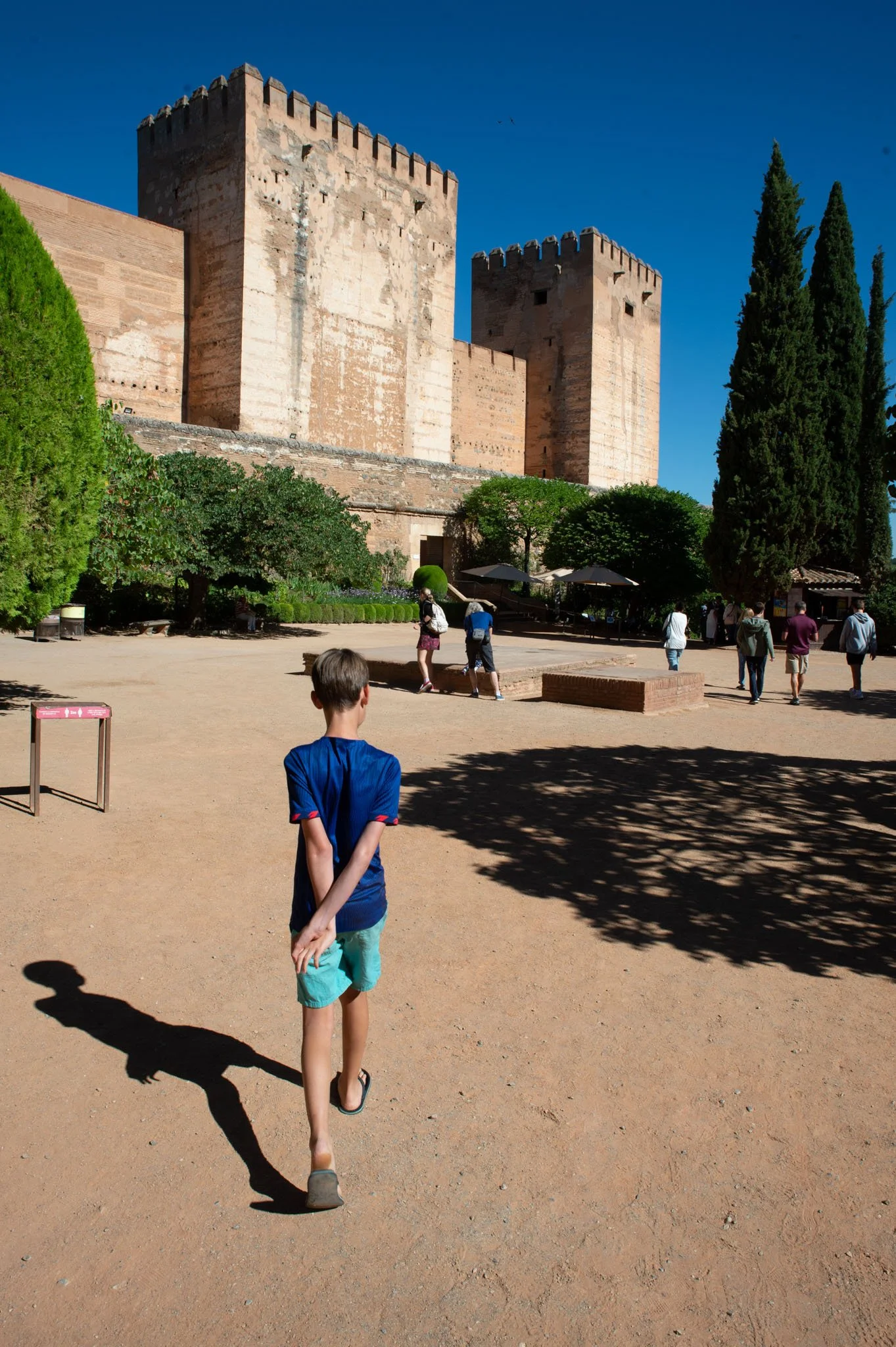 A young boy in a blue shirt and turquoise shorts walking on a dirt path with a shadow cast on the ground, towards an ancient castle with large stone towers, surrounded by greenery and a group of people.