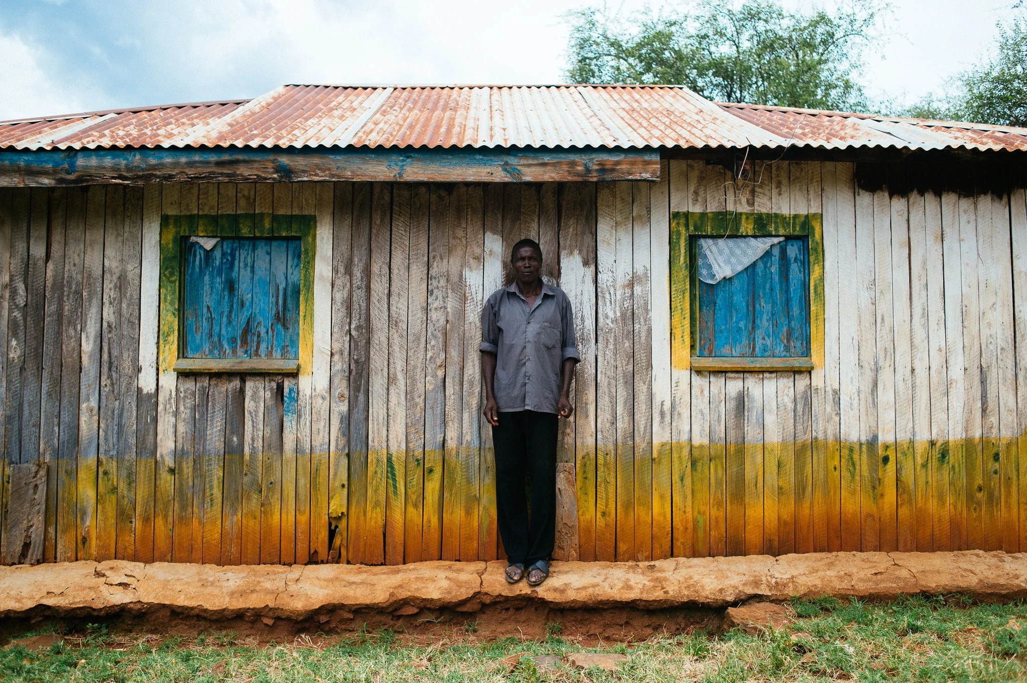 A man stands in front of a weathered wooden house with painted blue window frames and a corrugated metal roof. The house is set on a dirt ground with some green grass at the bottom of the image. Kenya