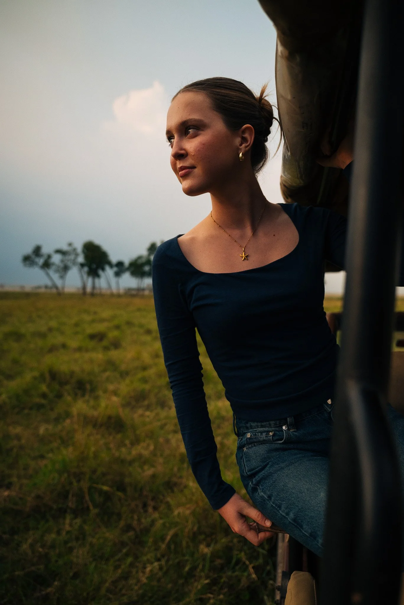 A woman leaning against a bus with a grassy field and trees in the background during the evening.