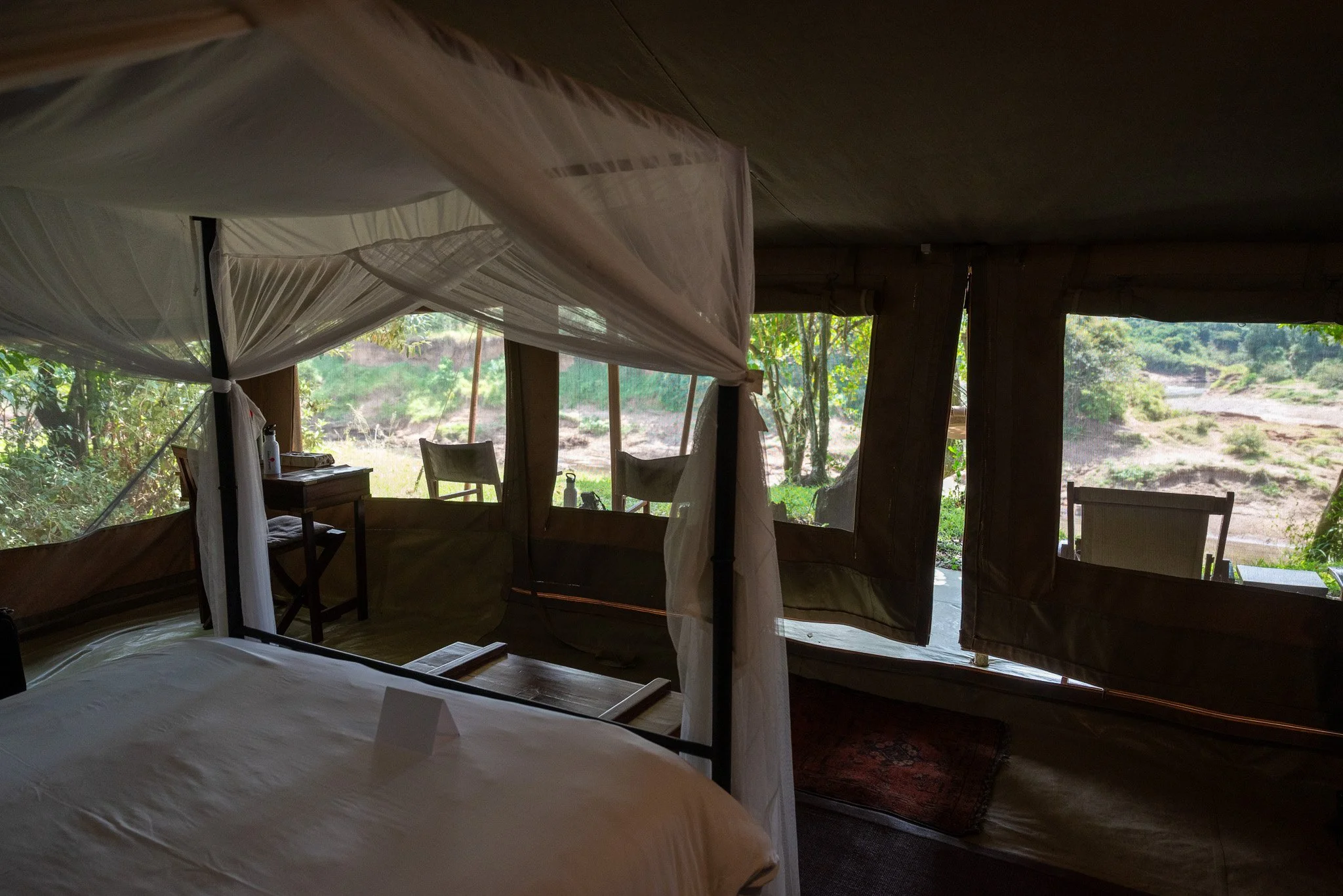 Interior of a rustic cabin bedroom with a bed and large open windows that show lush greenery and a river outside.