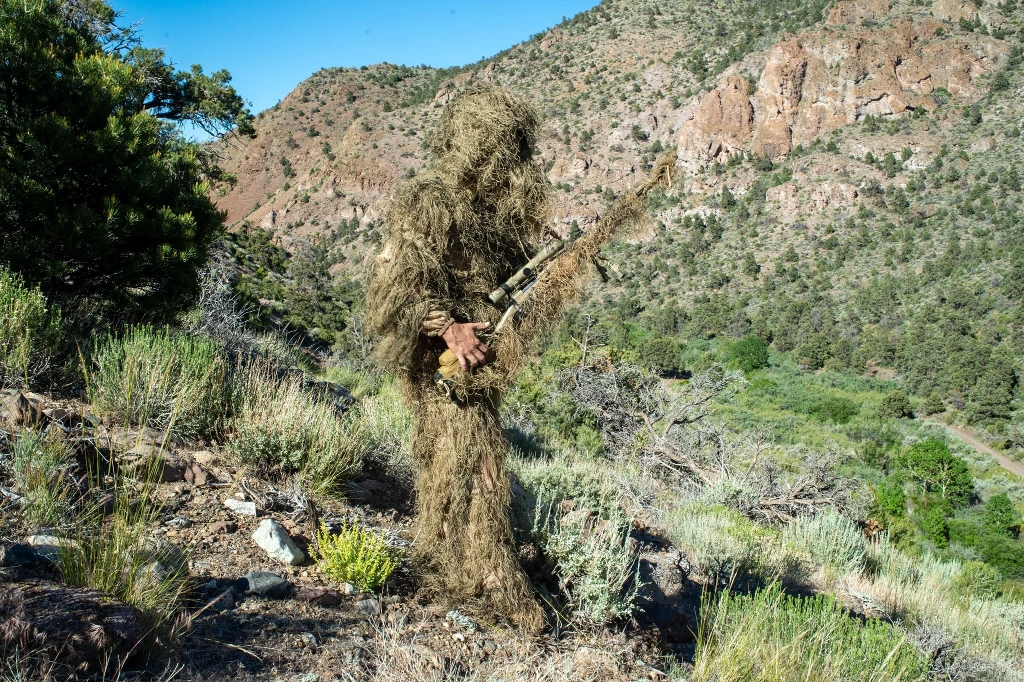 A person dressed in a ghillie suit holding a sniper rifle, standing on a rocky hillside in a mountainous area with green shrubs and trees.