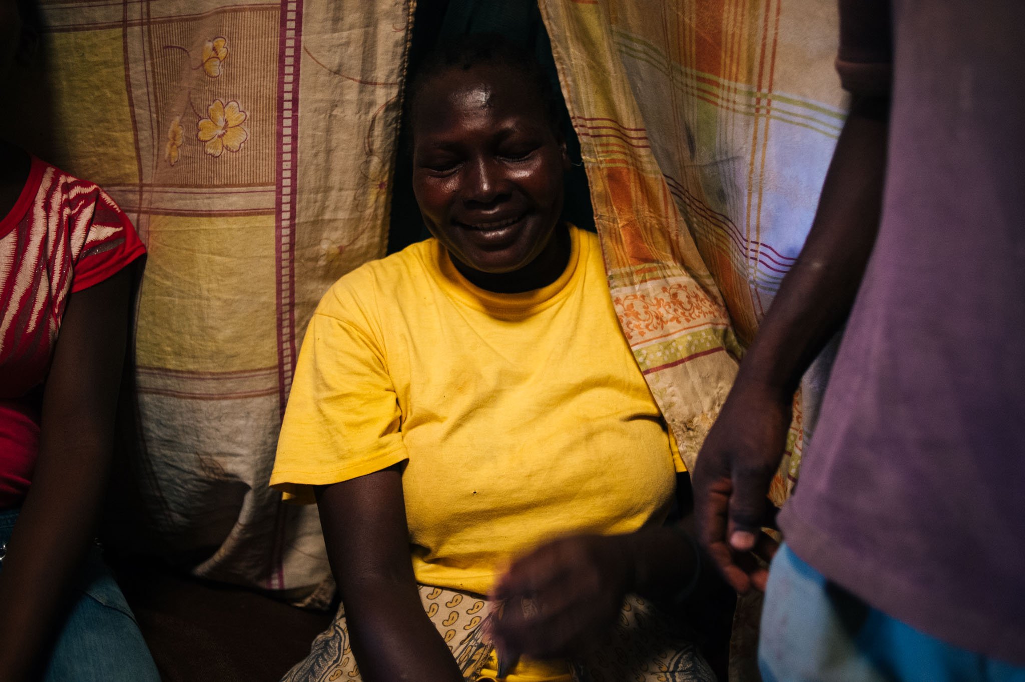 A woman in a yellow shirt sitting amidst a makeshift setting with fabric draped overhead, her face shows emotion, possibly tears of joy or sorrow, as another person stands nearby.