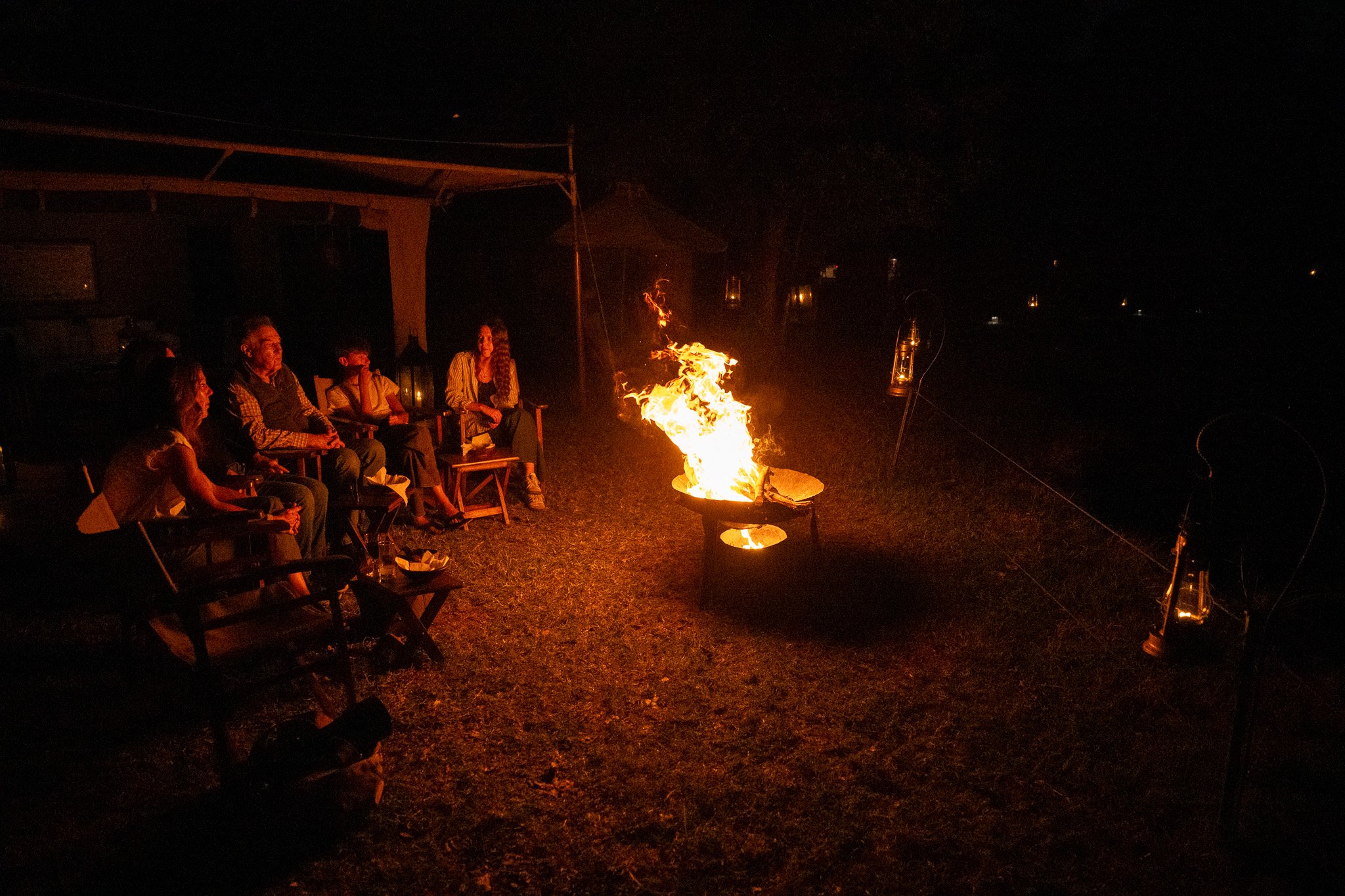 A group of people sitting around a campfire at night, with lanterns hanging in the background.