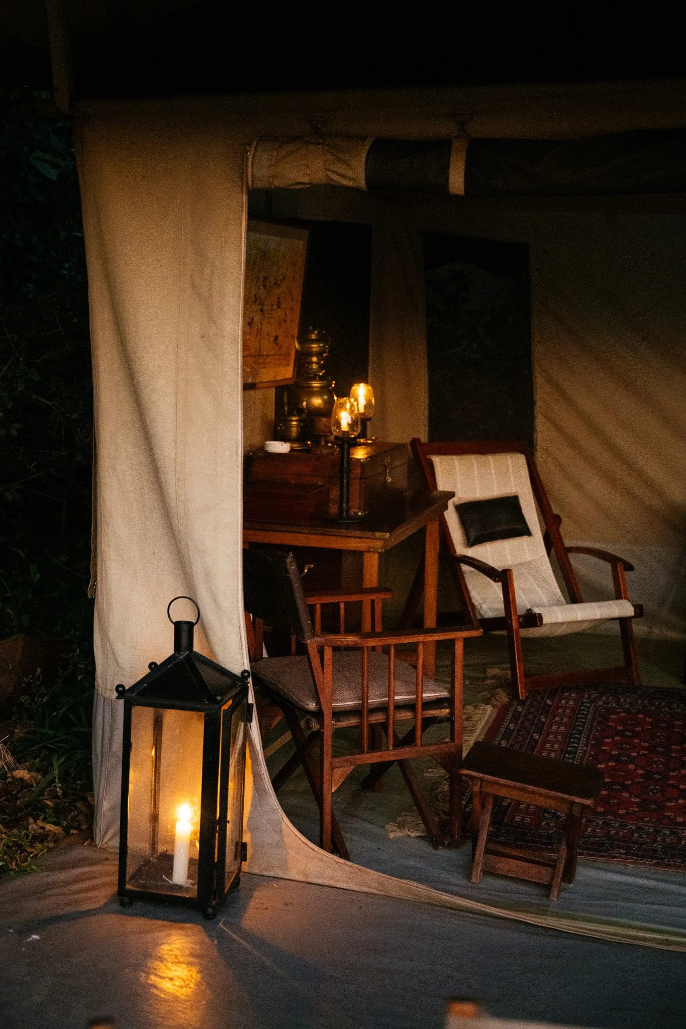 An outdoor scene at night with a patio setup. There are wooden chairs and a small table with a vintage lamp, a lantern with a candle, and some decorative items. The area is partially covered by a canopy or tent with fabric sides, and some greenery is