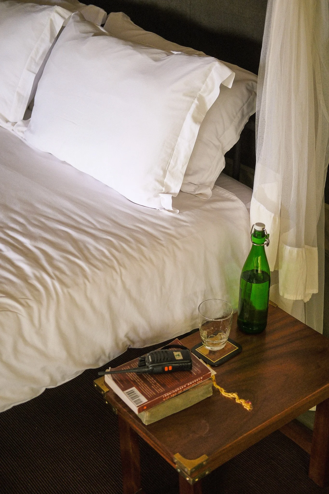 A bedside table with a green glass bottle, a glass of water, a closed book, a remote control, and a notebook. A white bed with pillows and a white net curtain are in the background.