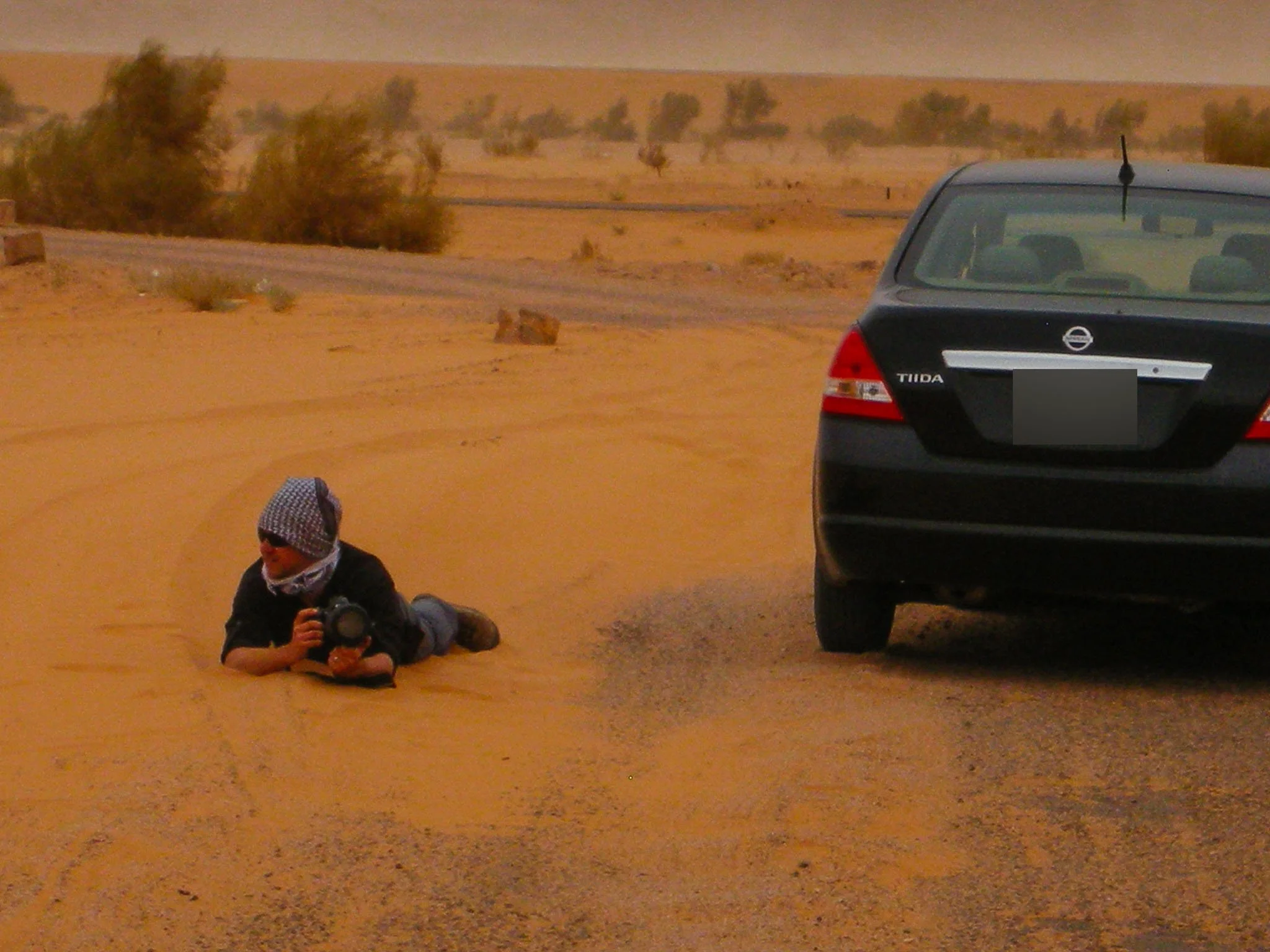 A person lying on the sand next to a black car, holding a camera, in a desert landscape with sparse bushes and a winding road in the background.