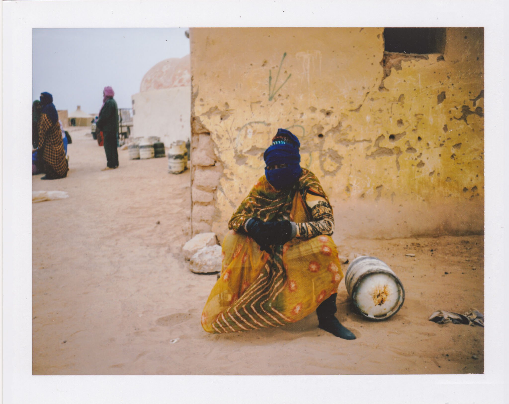 Person wearing colorful traditional clothing and a blue head covering, sitting on the ground next to a barrel, in front of a yellow, weathered wall in a desert-like setting.