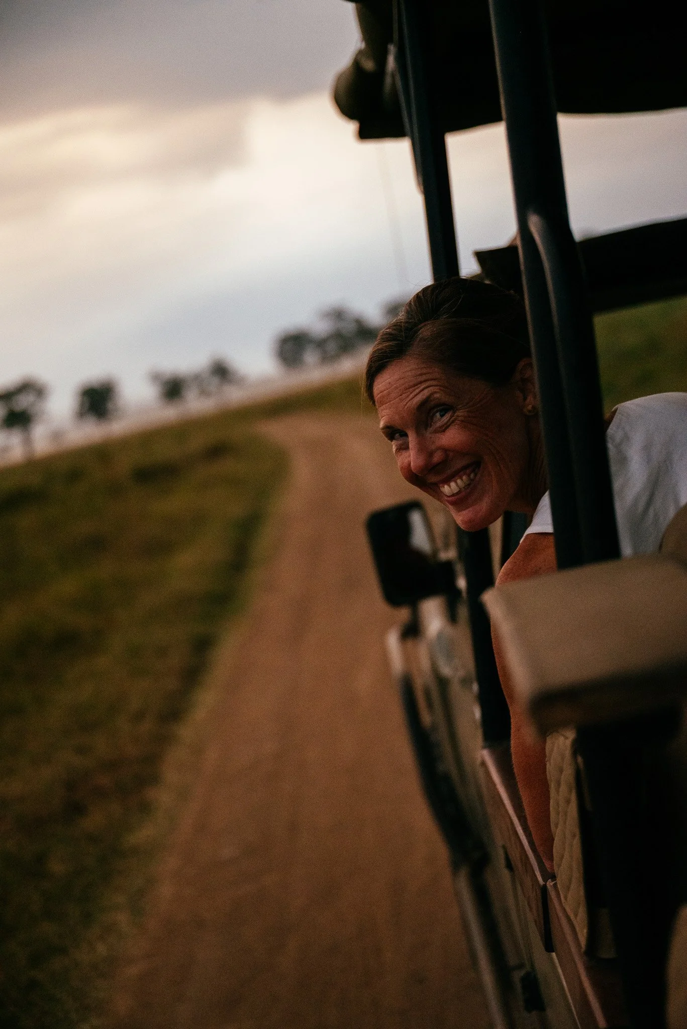 A woman smiling and looking out from a golf cart in a rural setting during sunset or dusk, with trees in the background.