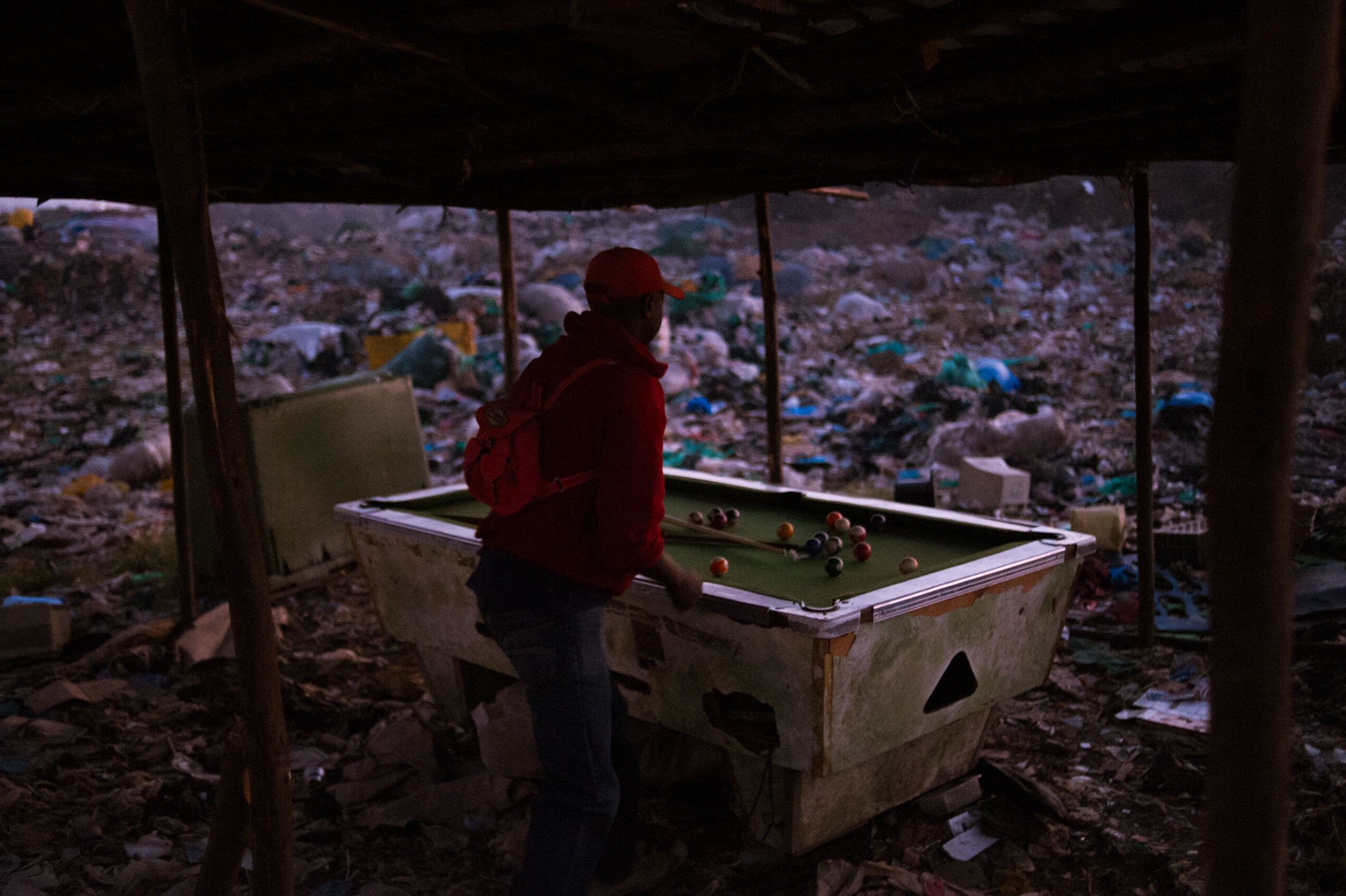 A person playing pool inside a makeshift shelter in a trash-filled area, with plastic waste and debris covering the ground. Dandora, Kenya