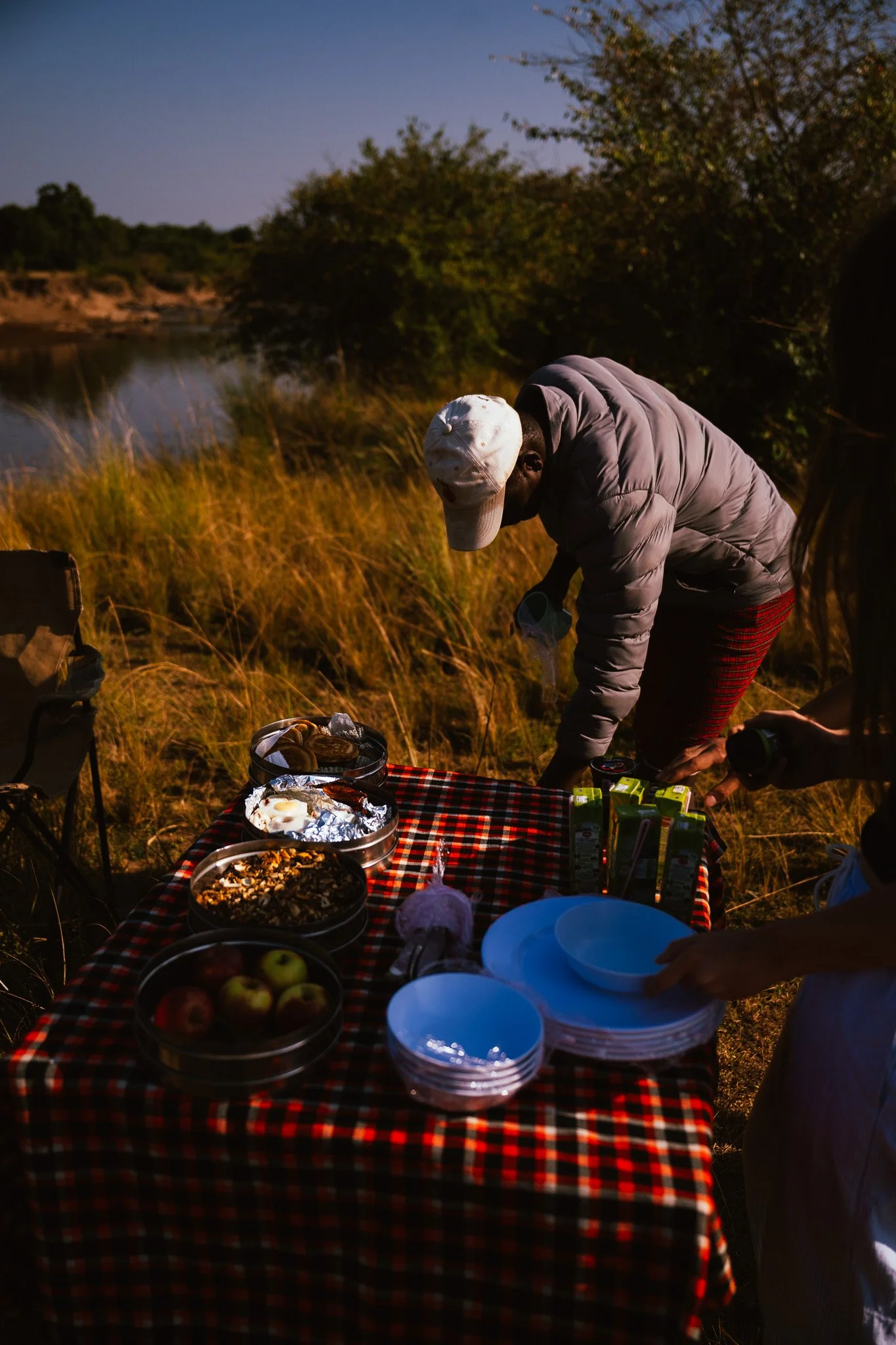 People having a picnic outdoors near a river during sunset, with food and drinks on a checkered tablecloth.