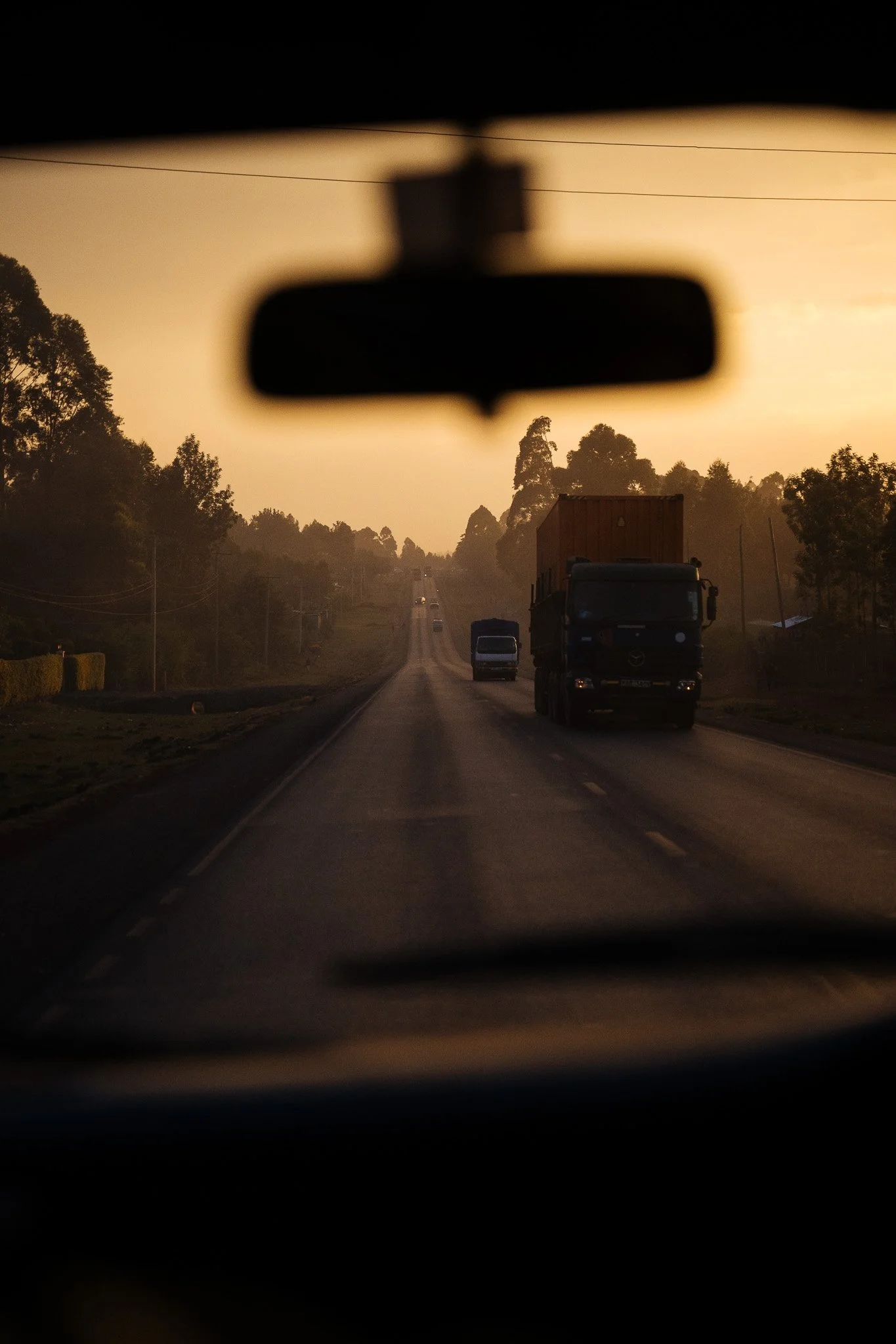 A view through a car's rearview mirror showing a highway with trucks and trees on both sides during sunset. Turbo, Kenya