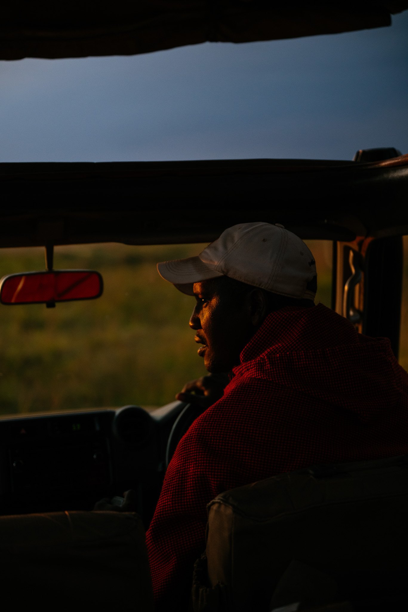 A person wearing a white cap and glasses, sitting in a vehicle, looking out the window at sunset or sunrise, with a landscape visible outside.