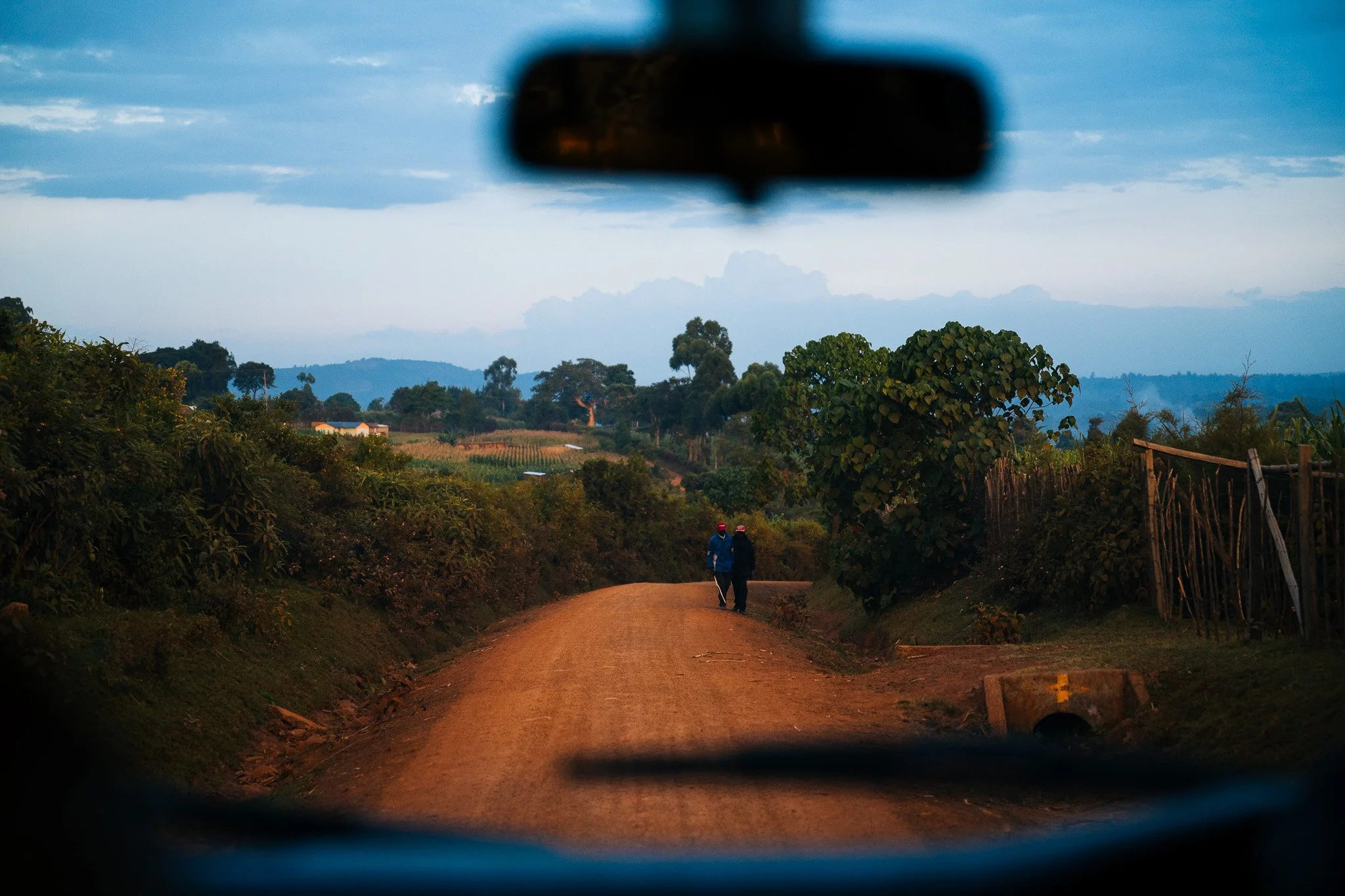 View through a car's rearview mirror showing a dirt road, two people walking, and a rural landscape with trees and hills under a blue sky with clouds. Kenya