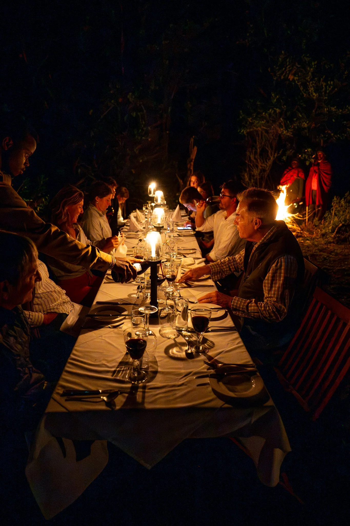 People gathered around a long dinner table outdoors at night, illuminated by candles, with trees in the background and a fire with individuals in traditional clothing nearby.