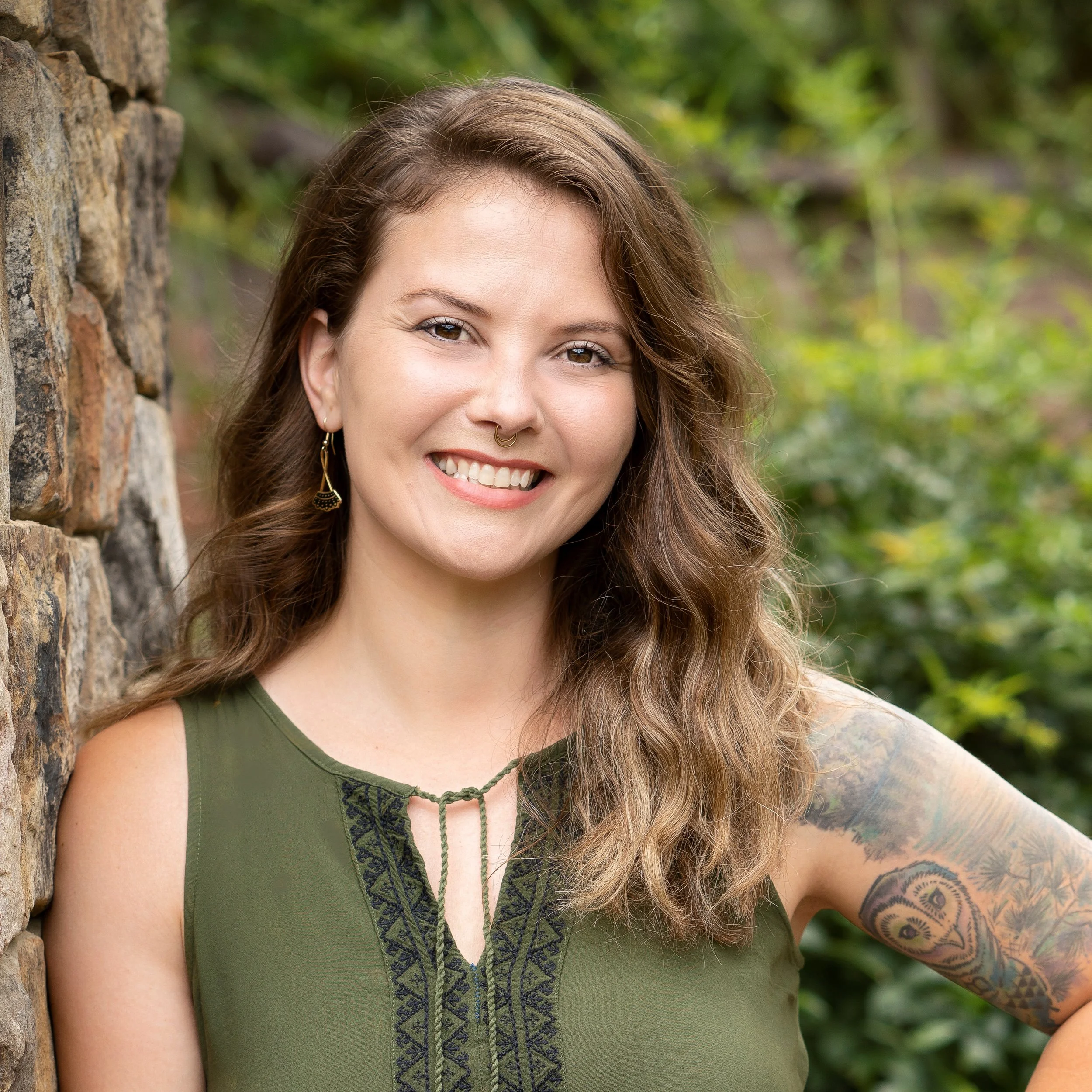 Portrait of a smiling woman with long wavy hair, jewelry, and a tattoo, standing outdoors near a stone wall with greenery in the background.