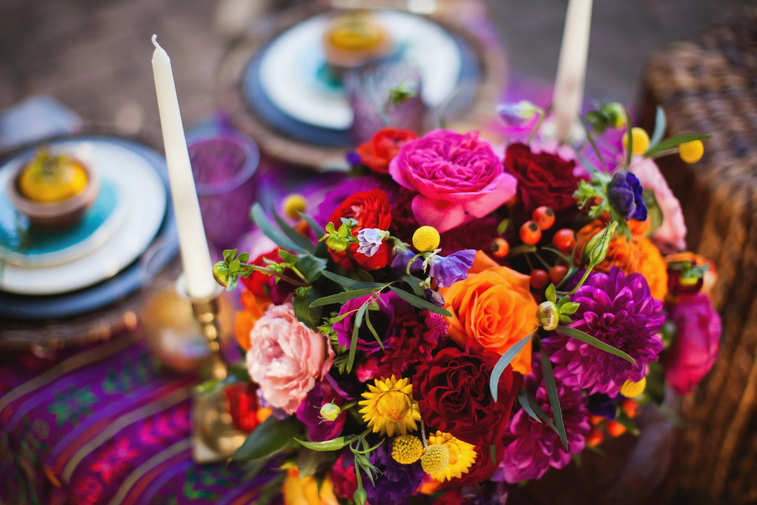 A colorful bouquet of pink, orange, purple, yellow, and red flowers with green leaves on a table, with two white candles and decorative plates in the background.