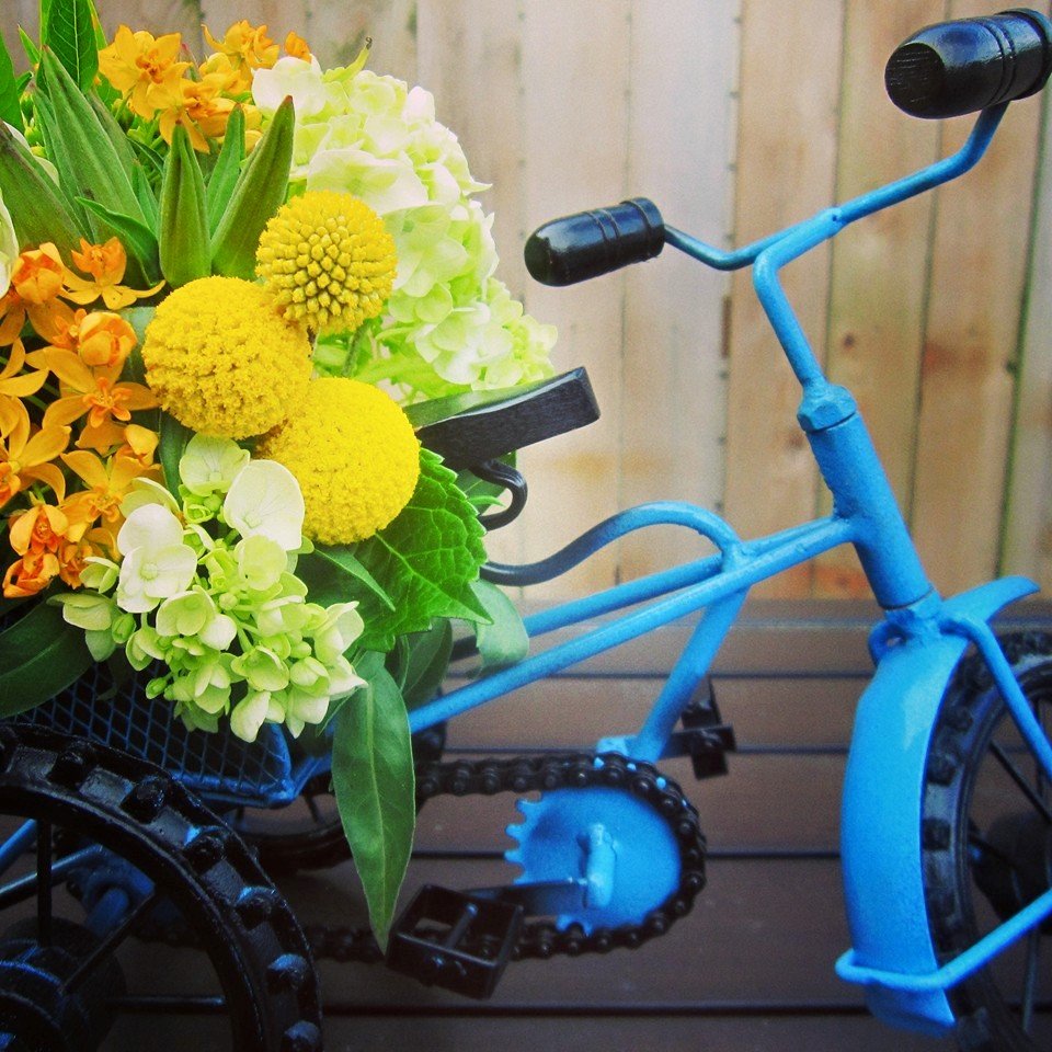 A blue miniature bicycle with a basket of yellow, white, and orange flowers on it, set against a wooden fence background.