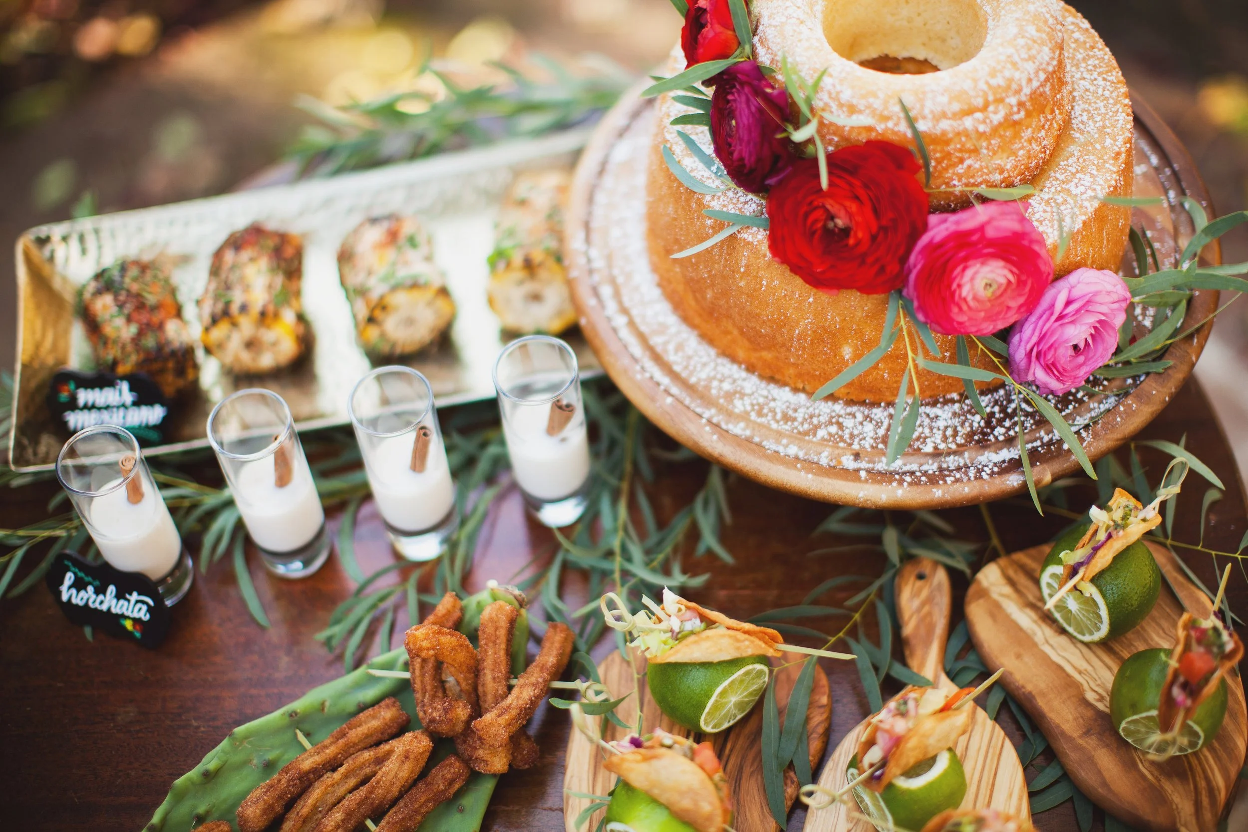 A table with a bundt cake decorated with pink and red flowers, small glasses of horchata, sushi rolls, fried churros, and fruit tacos garnished with lime slices and greenery.