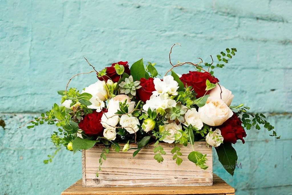 A wooden box filled with white, red, and pale pink flowers with green foliage, set against a turquoise brick wall background.